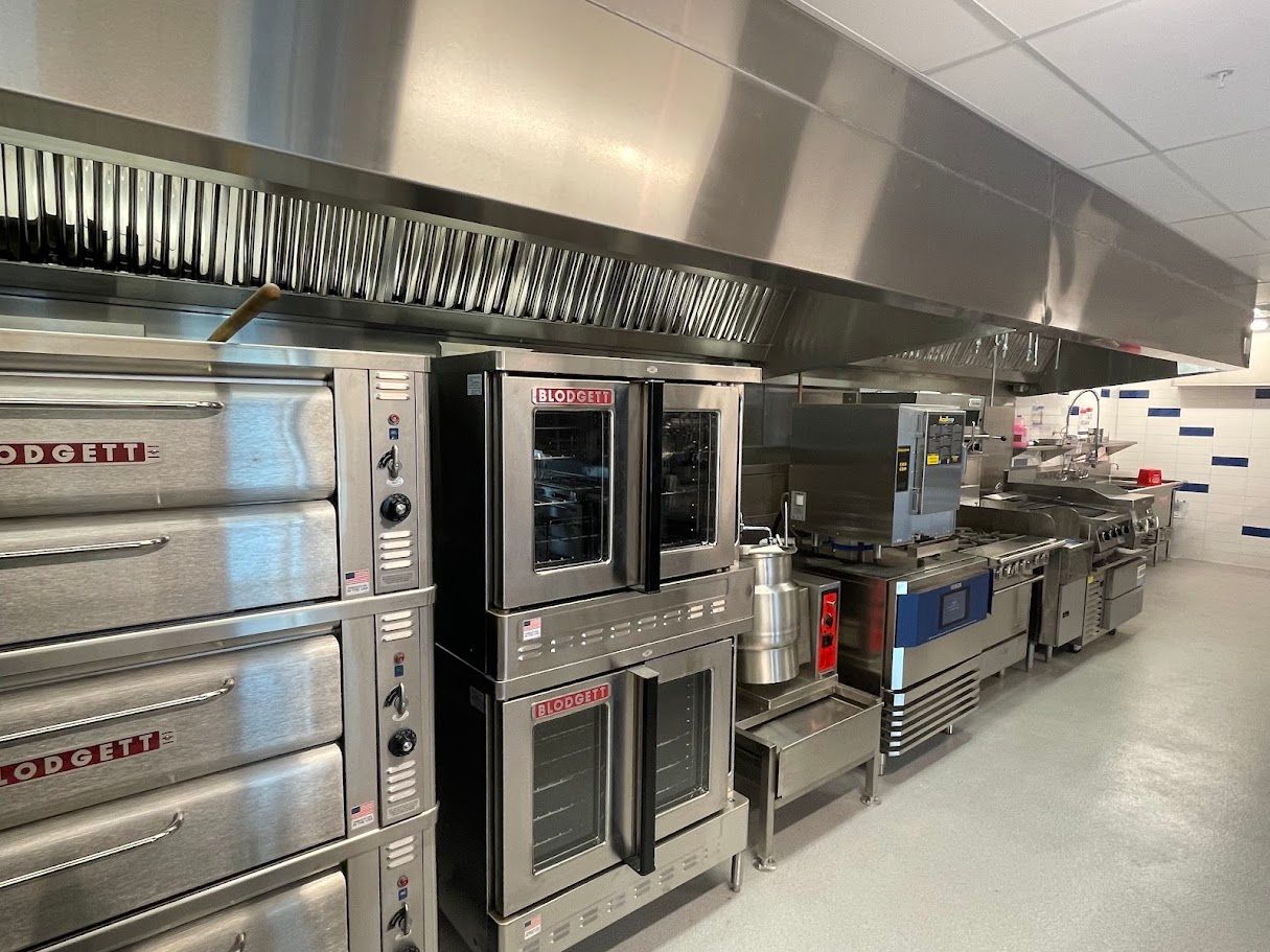 A row of stainless steel commercial kitchen appliances, including stacked ovens, under a large metal ventilation hood.