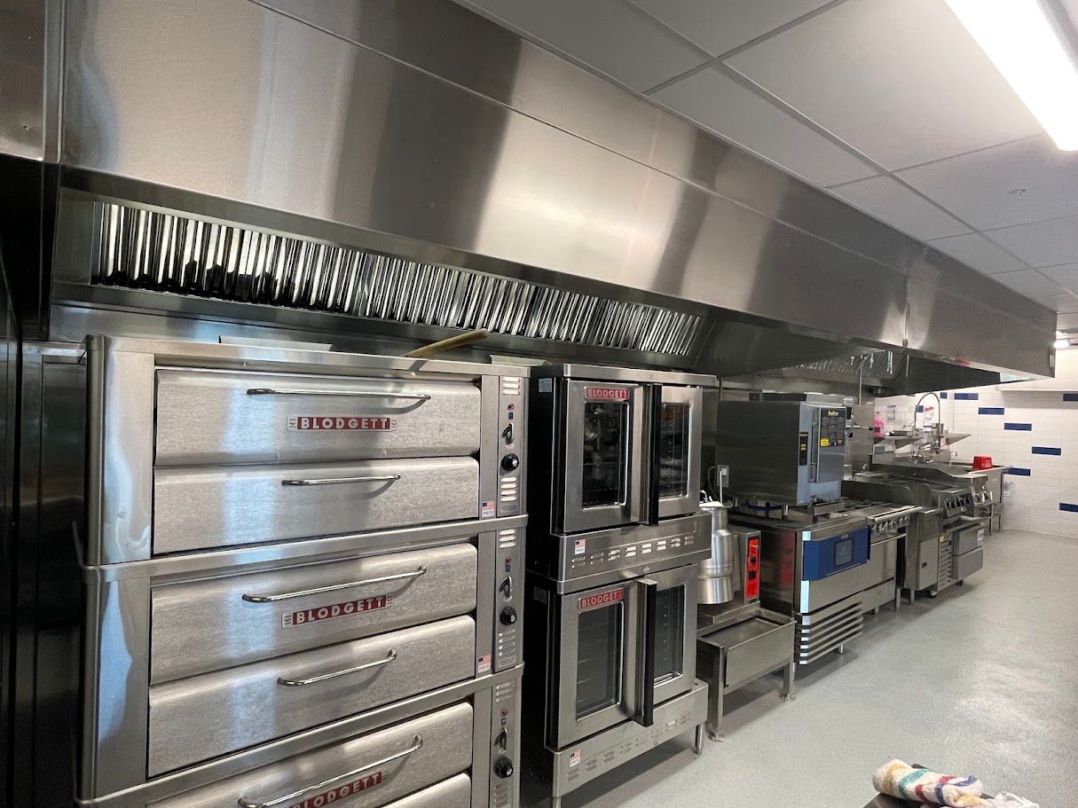 A professional kitchen featuring stainless steel pizza ovens, convection ovens, and an overhead ventilation hood.