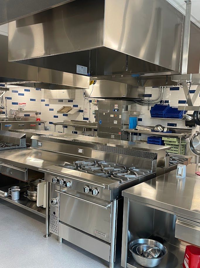 A professional kitchen with a large stainless steel range, oven, and vent hood, set against tiled walls with blue accents.
