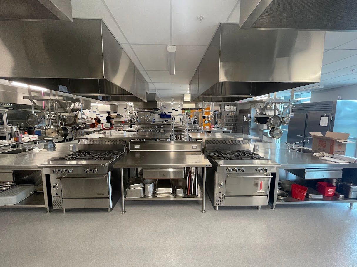 A clean, industrial commercial kitchen with rows of stainless steel stoves, prep tables, and overhead ventilation hoods.