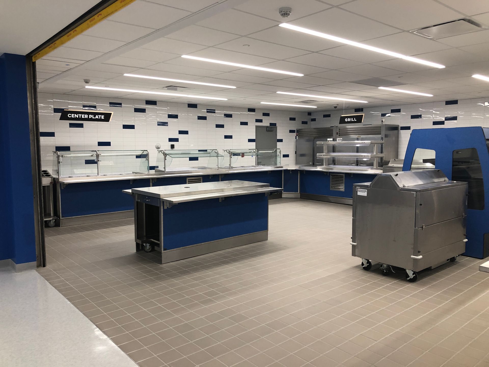 A modern cafeteria serving area with blue service counters, stainless steel food stations, and white tiled walls.