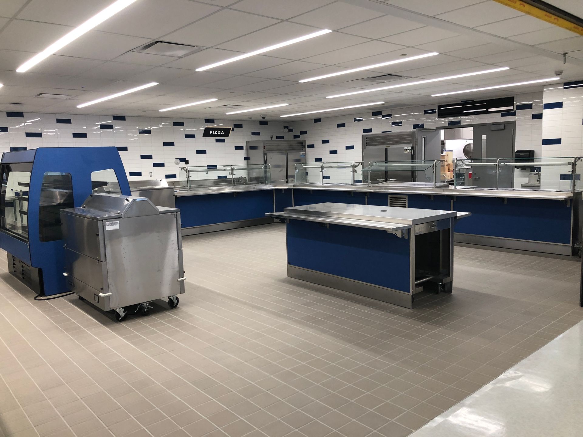 An empty commercial cafeteria serving line with stainless steel counters and blue base panels under bright lights.