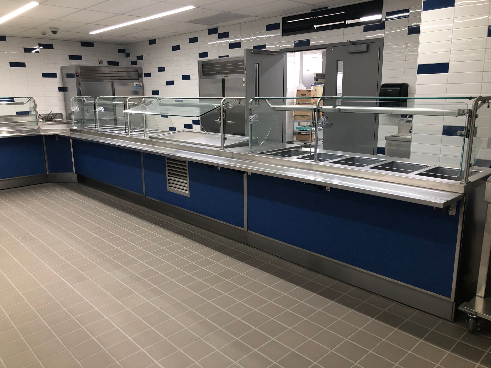 A long, empty cafeteria serving line with a stainless steel counter and glass sneeze guards in a tiled room.