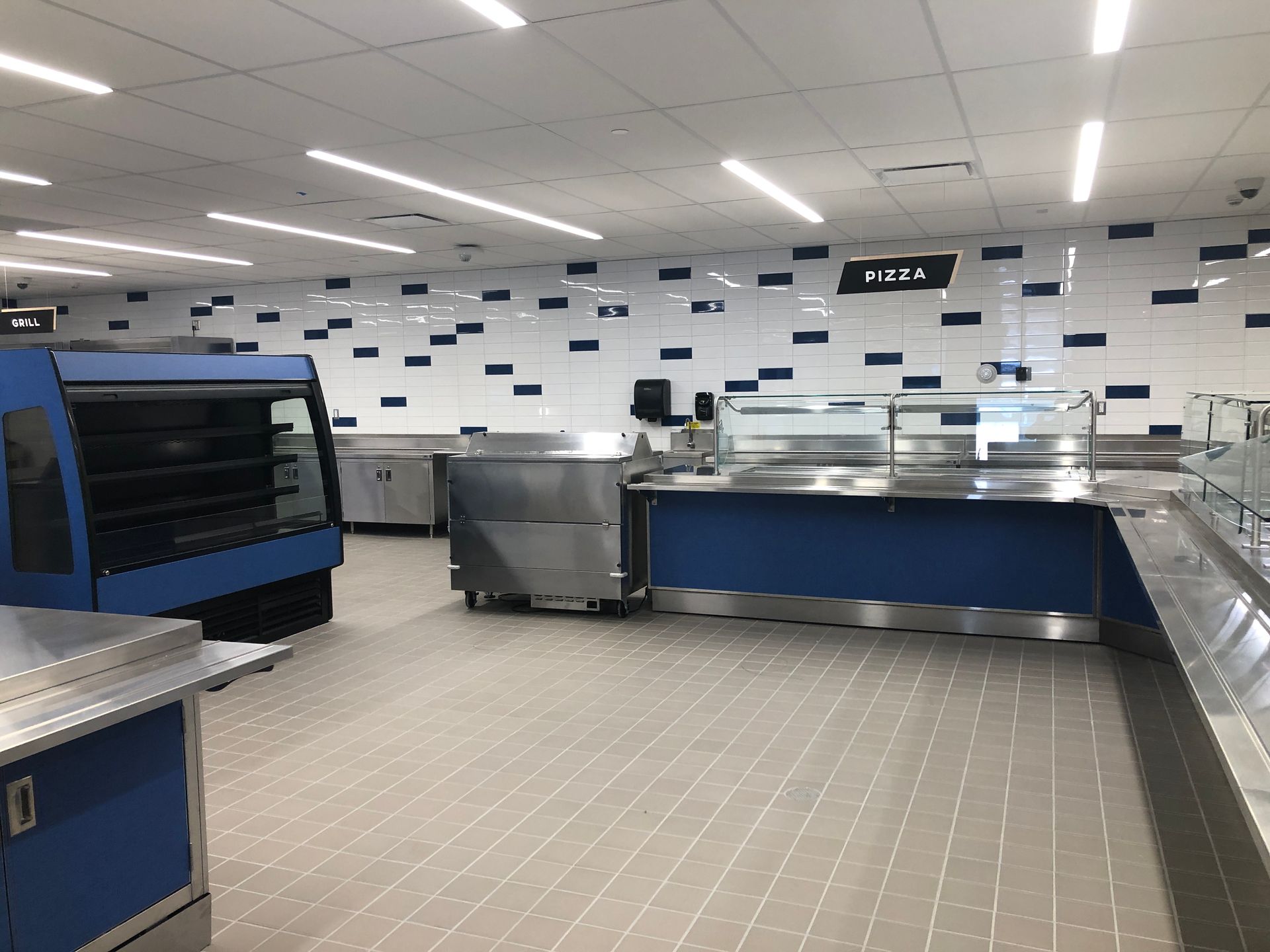 An empty cafeteria serving line with blue counters, stainless steel equipment, and white tiled walls with blue accents.