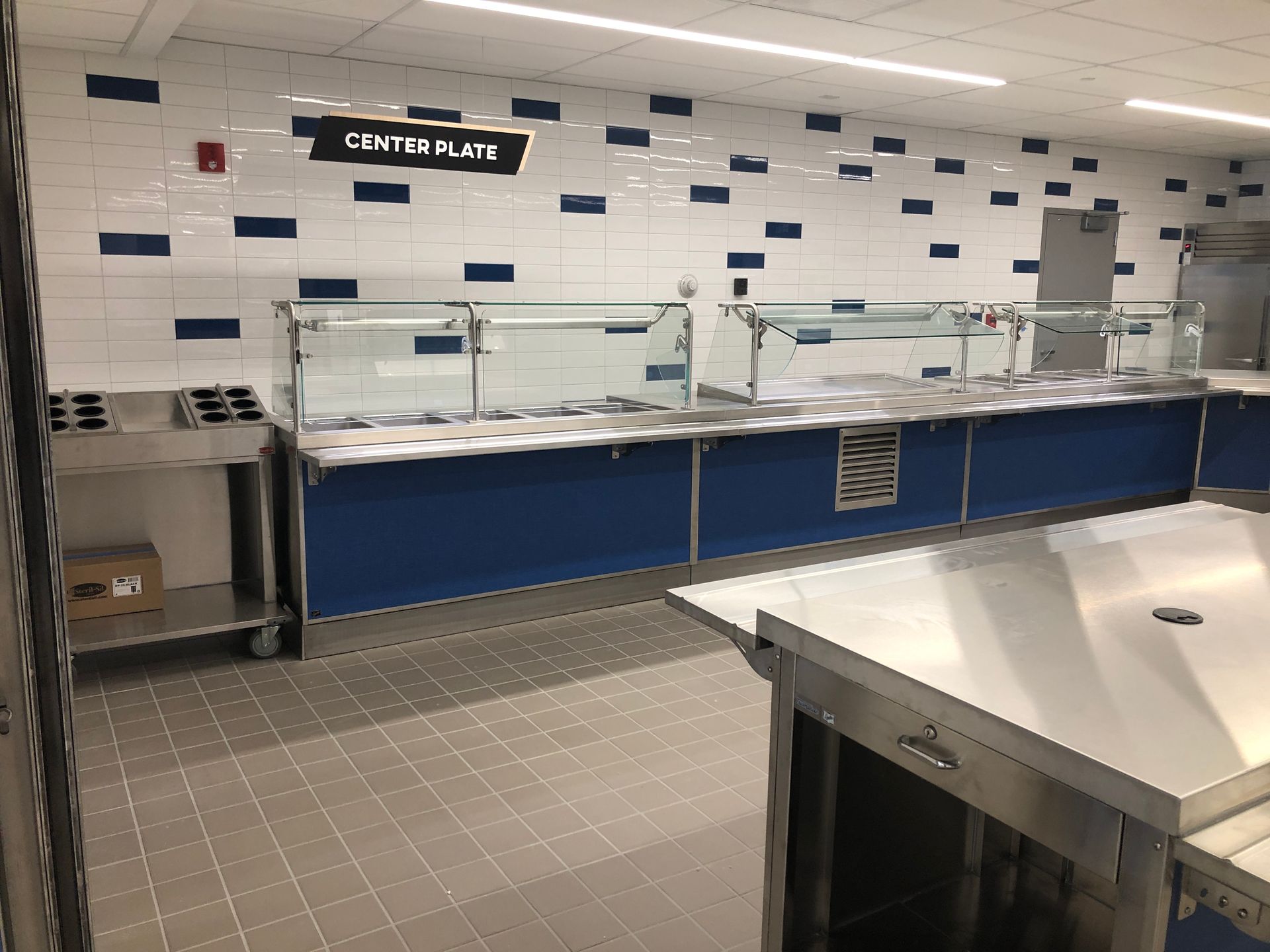 A modern cafeteria serving area with stainless steel counters, glass sneeze guards, and white tiled walls with blue accents.