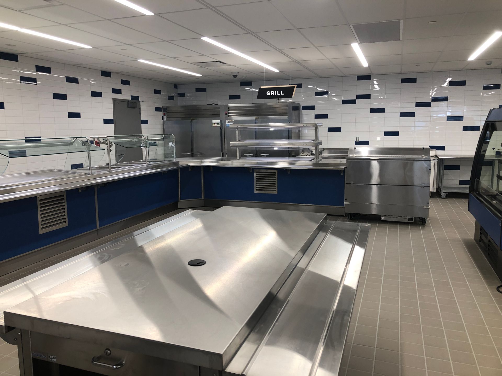 An empty cafeteria serving area featuring stainless steel counters, a tiled floor, and walls with black and white tiles.