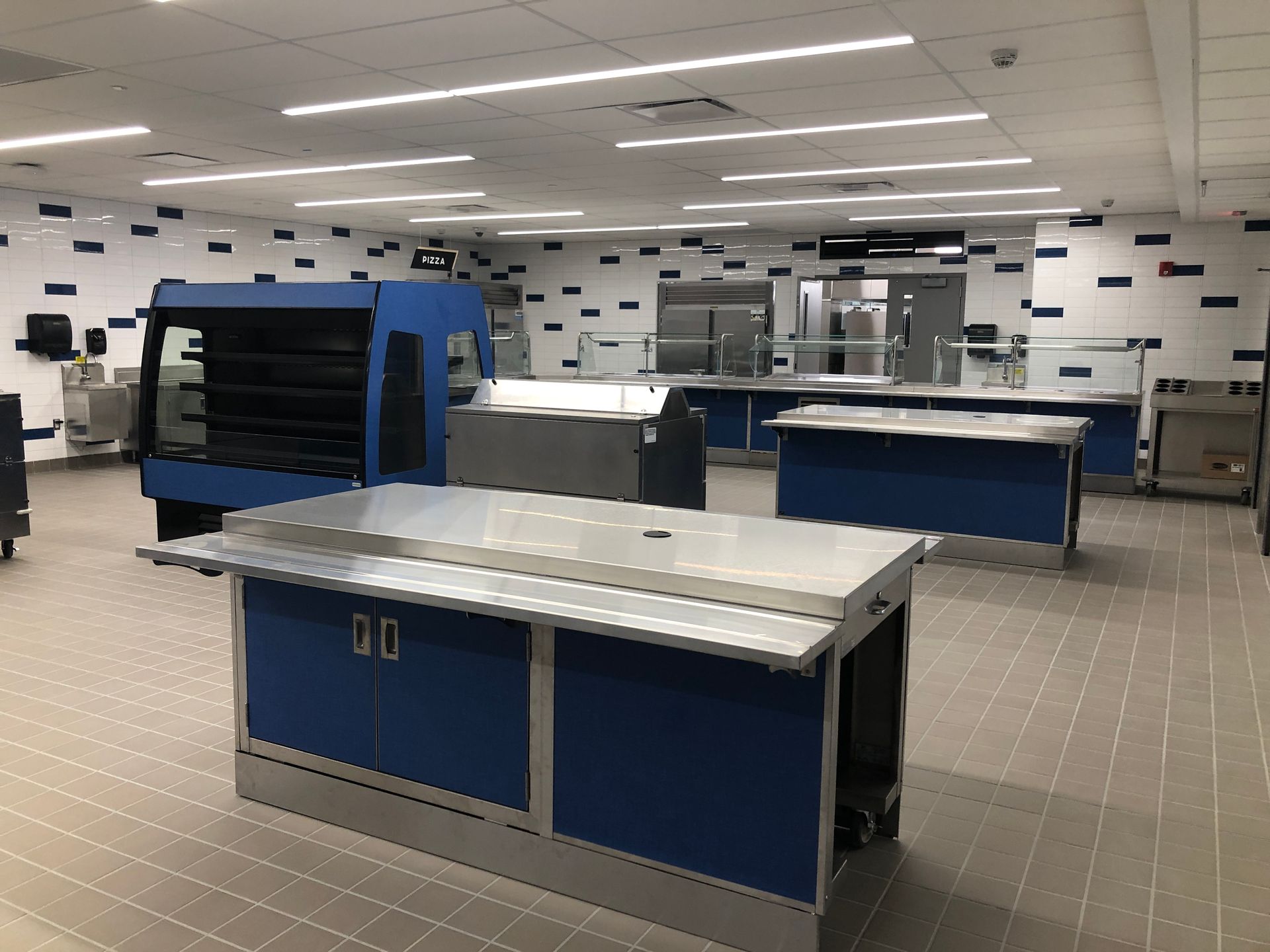 An empty cafeteria serving area featuring blue-paneled stainless steel food stations and tiled walls with blue accents.