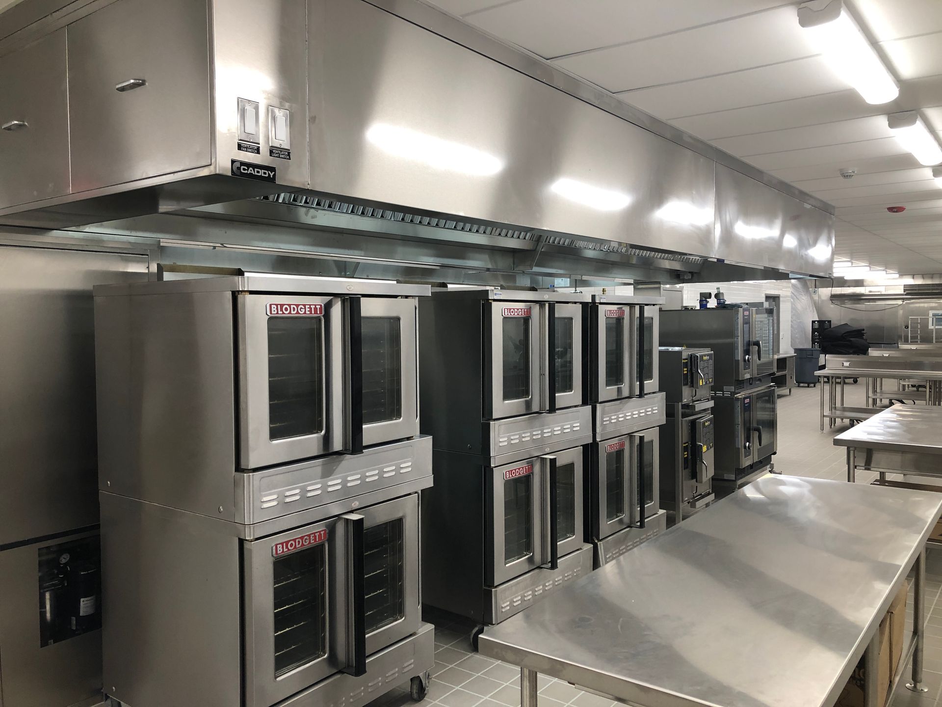 A row of industrial stainless steel ovens sits beneath a large kitchen ventilation hood in a commercial kitchen space.
