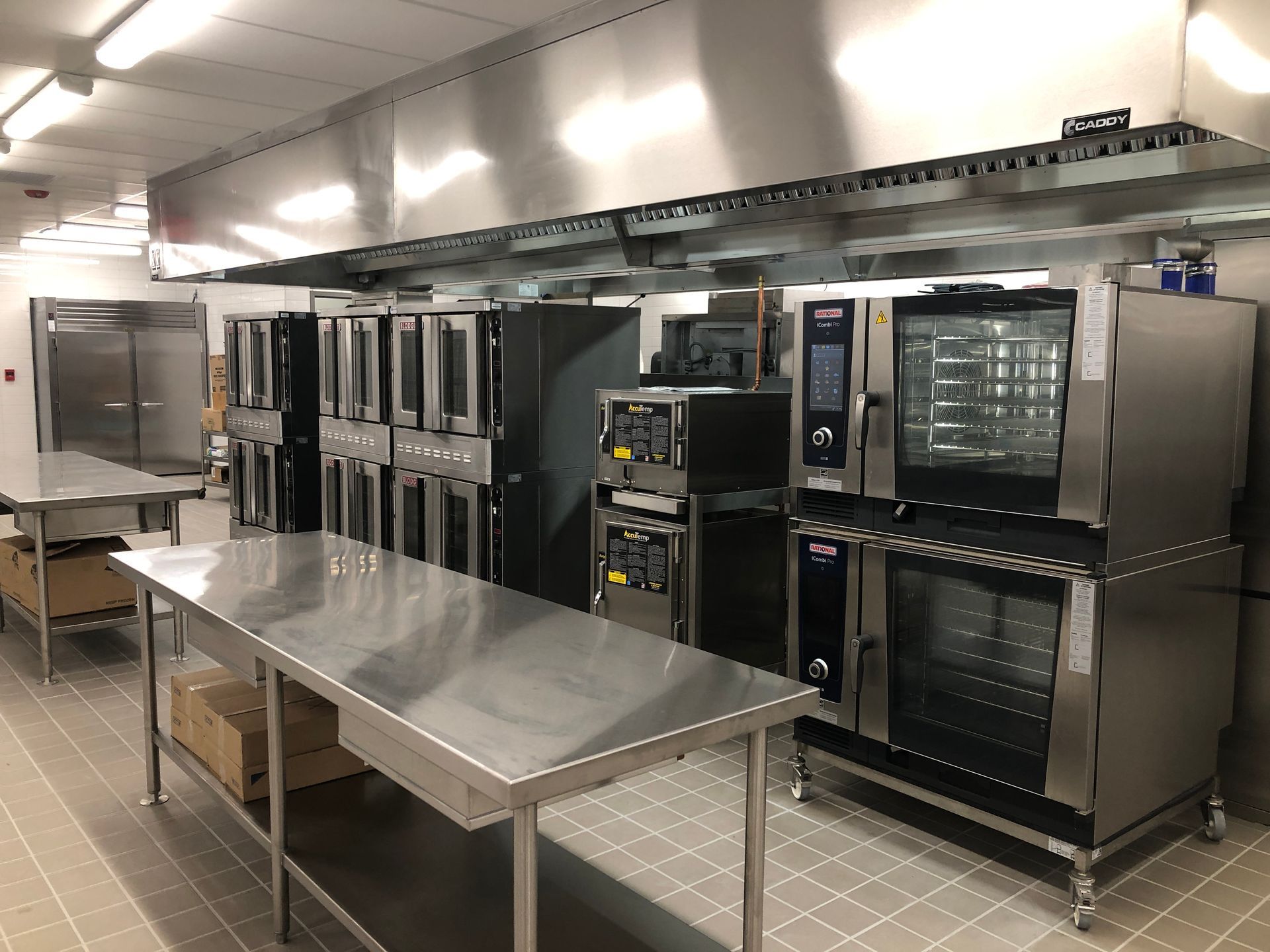A professional commercial kitchen with stainless steel prep tables, stacked convection ovens, and a large vent hood.