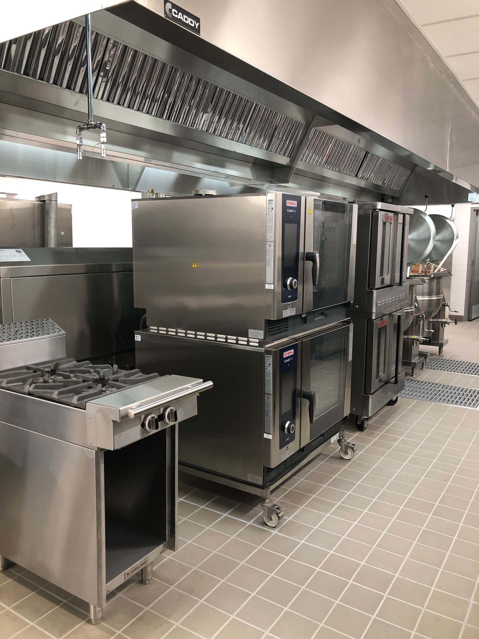 A row of stainless steel commercial kitchen appliances, including stoves and ovens, positioned under a large metal hood.