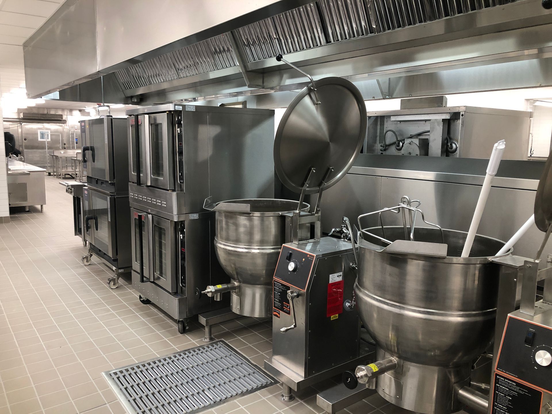 A professional kitchen featuring industrial stainless-steel ovens and large, upright cooking kettles under a range hood.