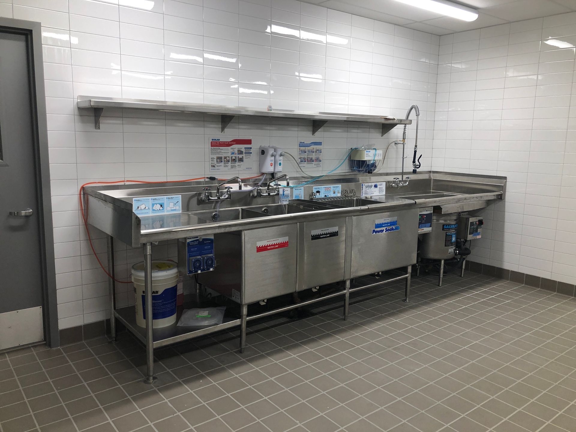 A stainless steel three-compartment commercial sink set against a white tiled wall in a clean, utilitarian kitchen.
