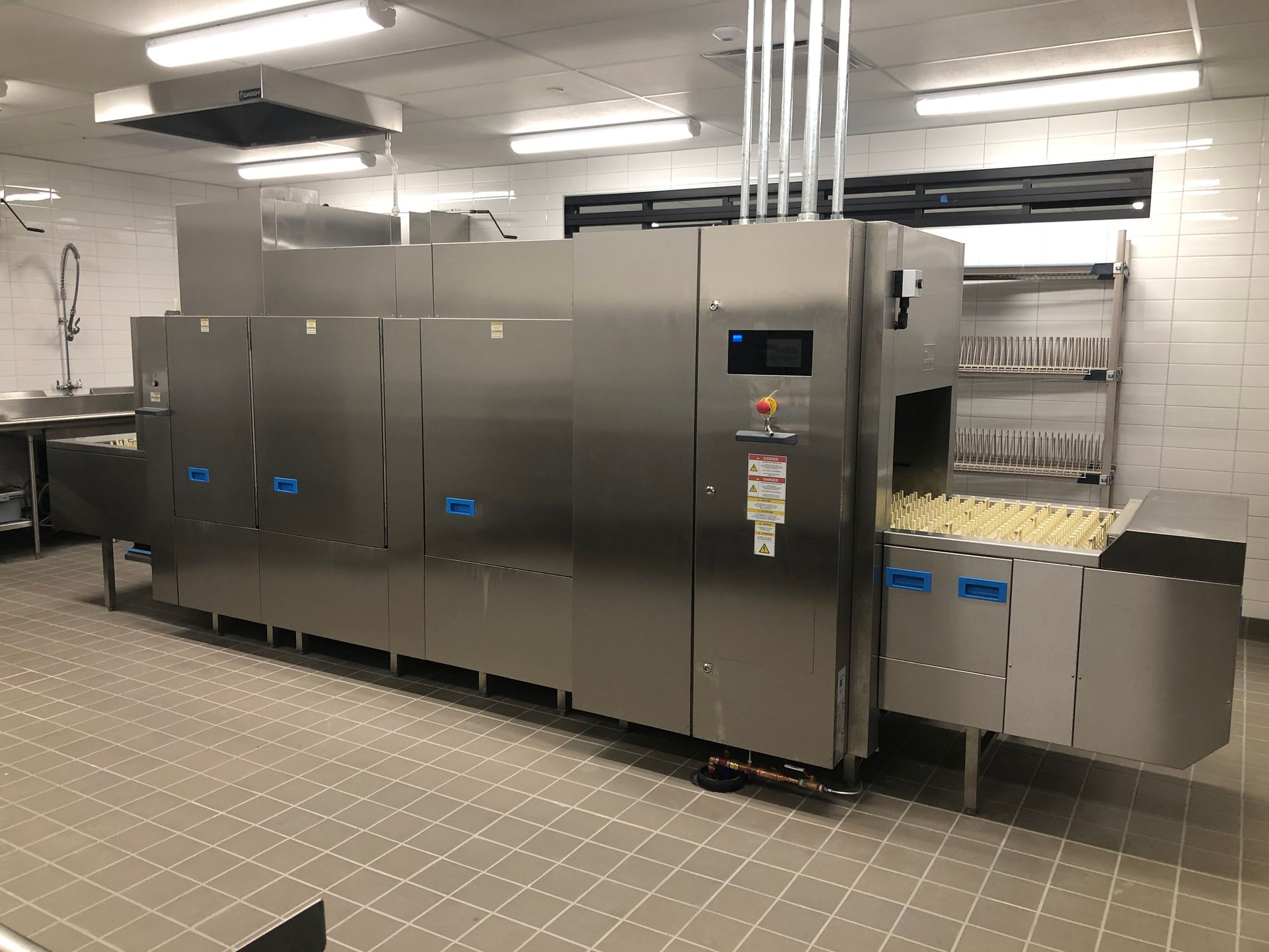 A large, industrial stainless steel conveyor dishwasher in a commercial kitchen with tiled floors and walls.