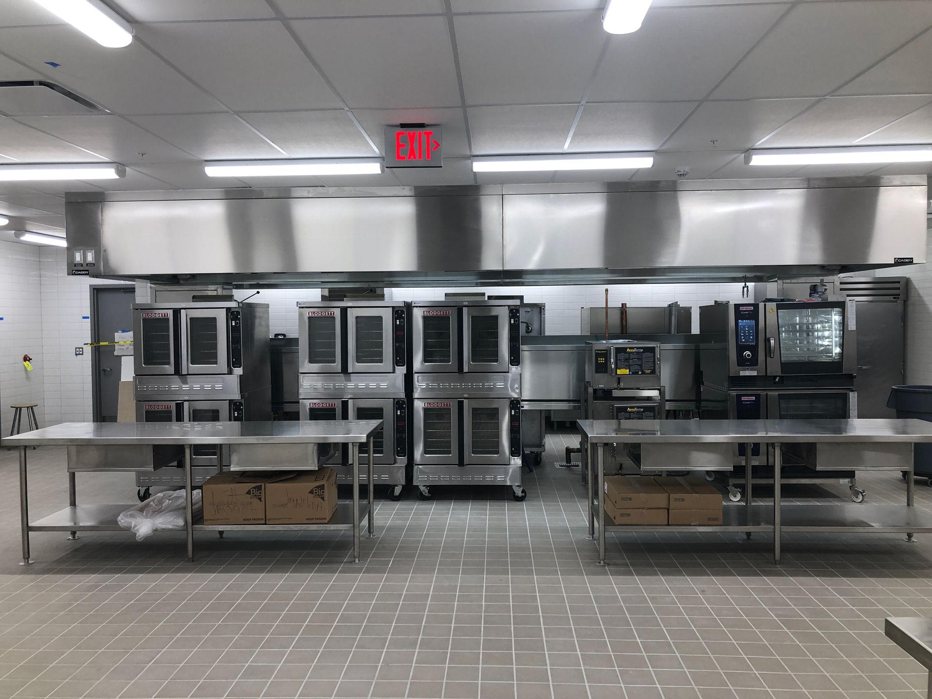 Commercial kitchen space featuring stainless steel ovens under a large vent hood and two metal work tables in front.