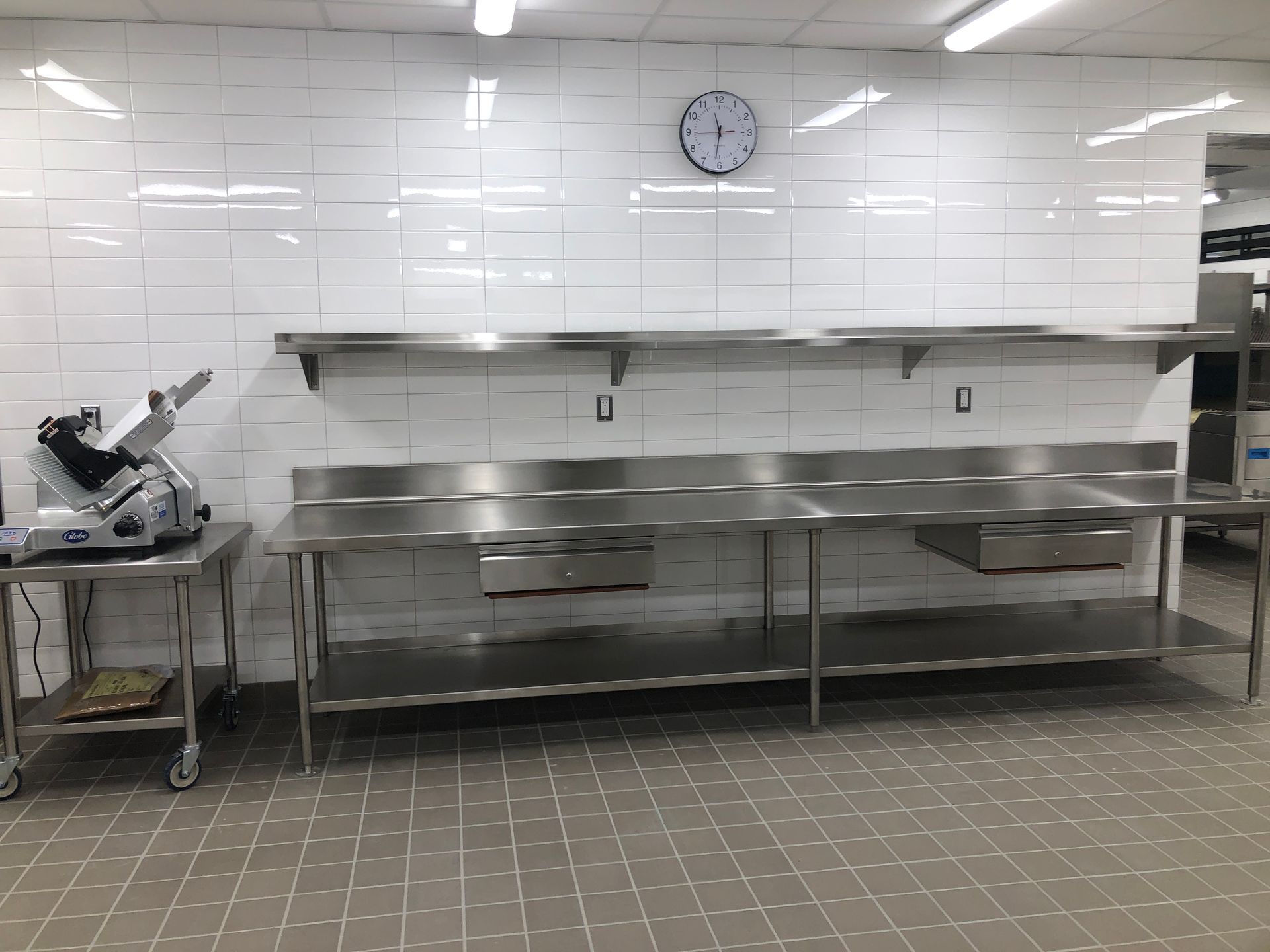 A commercial kitchen with a stainless steel prep table, a meat slicer on a side cart, and a wall clock against tiled walls.