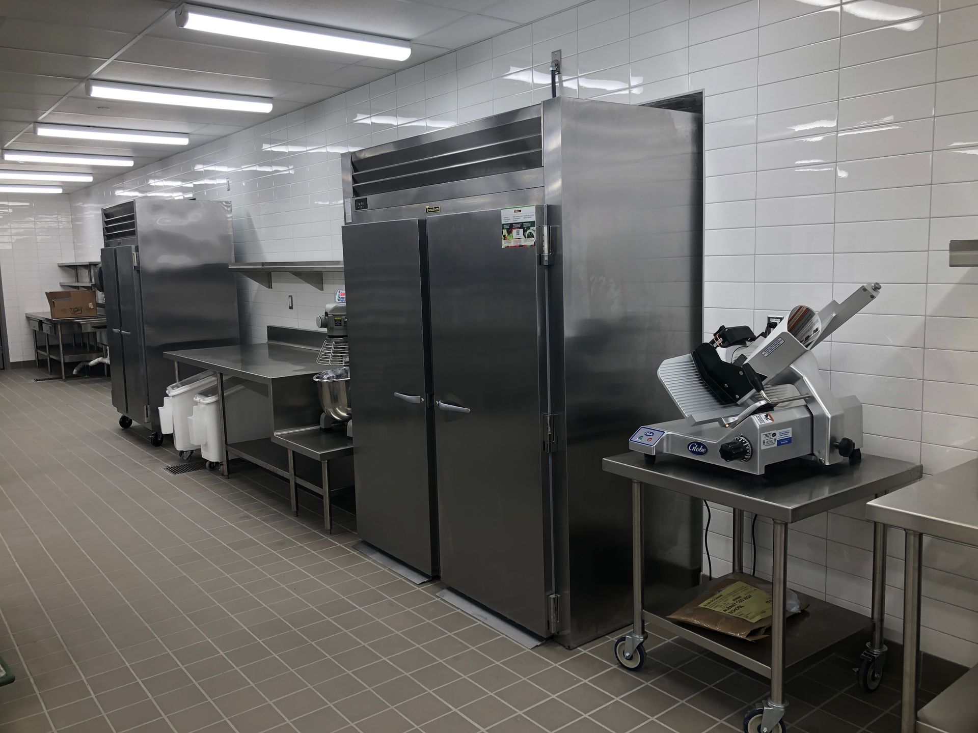 A commercial kitchen with stainless steel refrigerators and a meat slicer on a metal table, set against tiled walls.