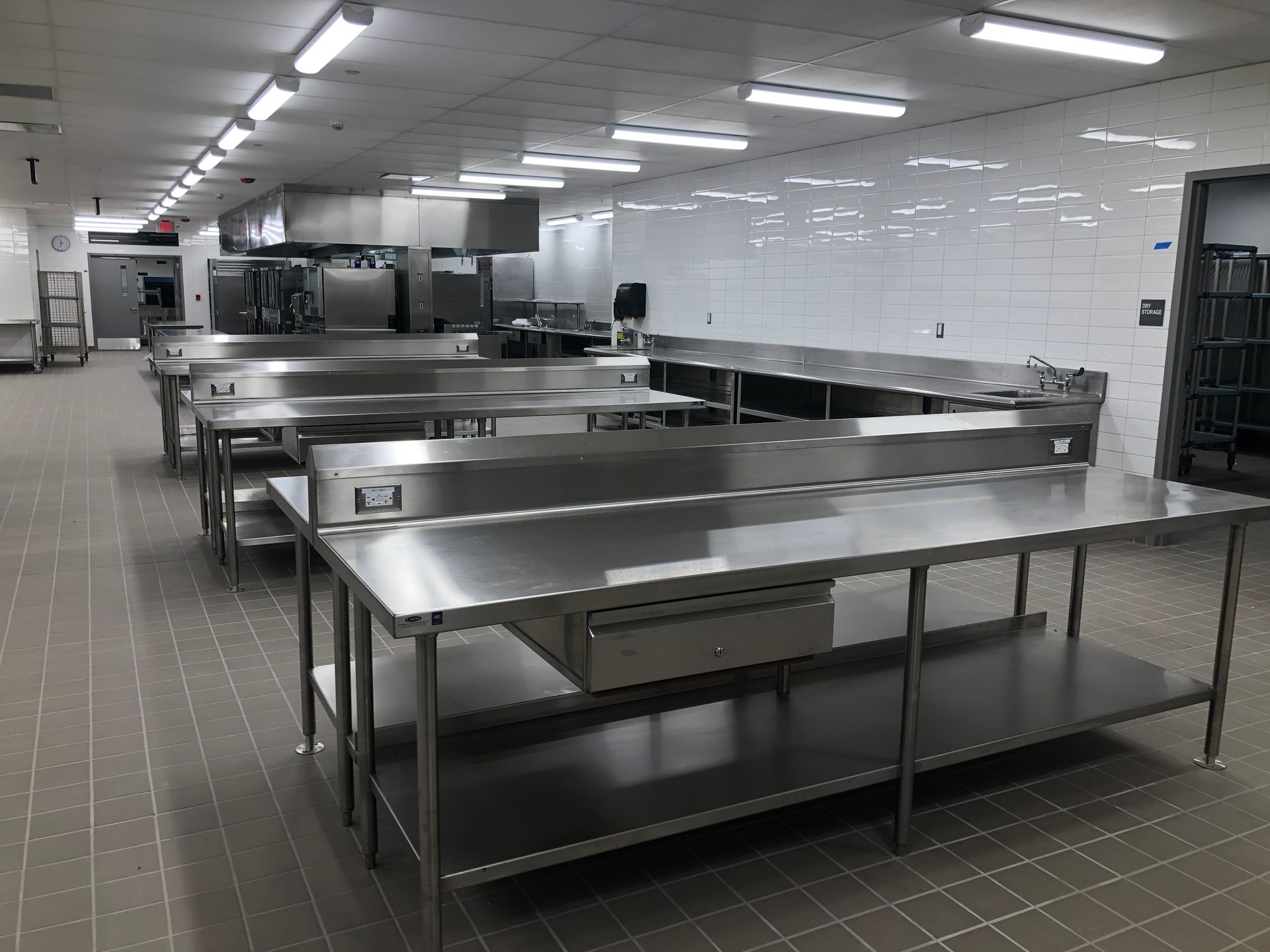A commercial kitchen featuring multiple stainless steel work tables arranged in rows against a tiled wall.