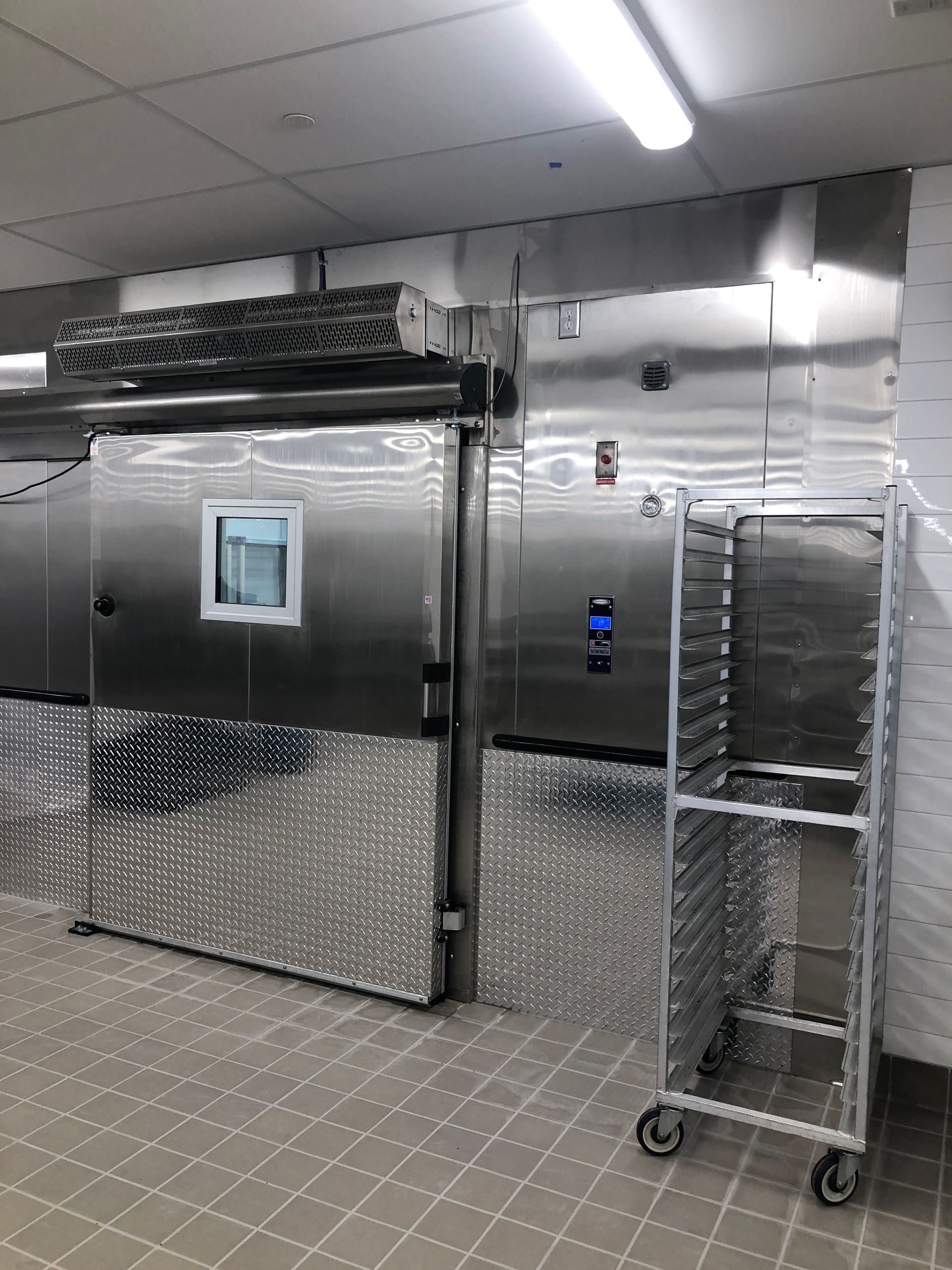 A stainless steel industrial walk-in freezer with a viewing window, digital control panel, and a rolling rack in front.