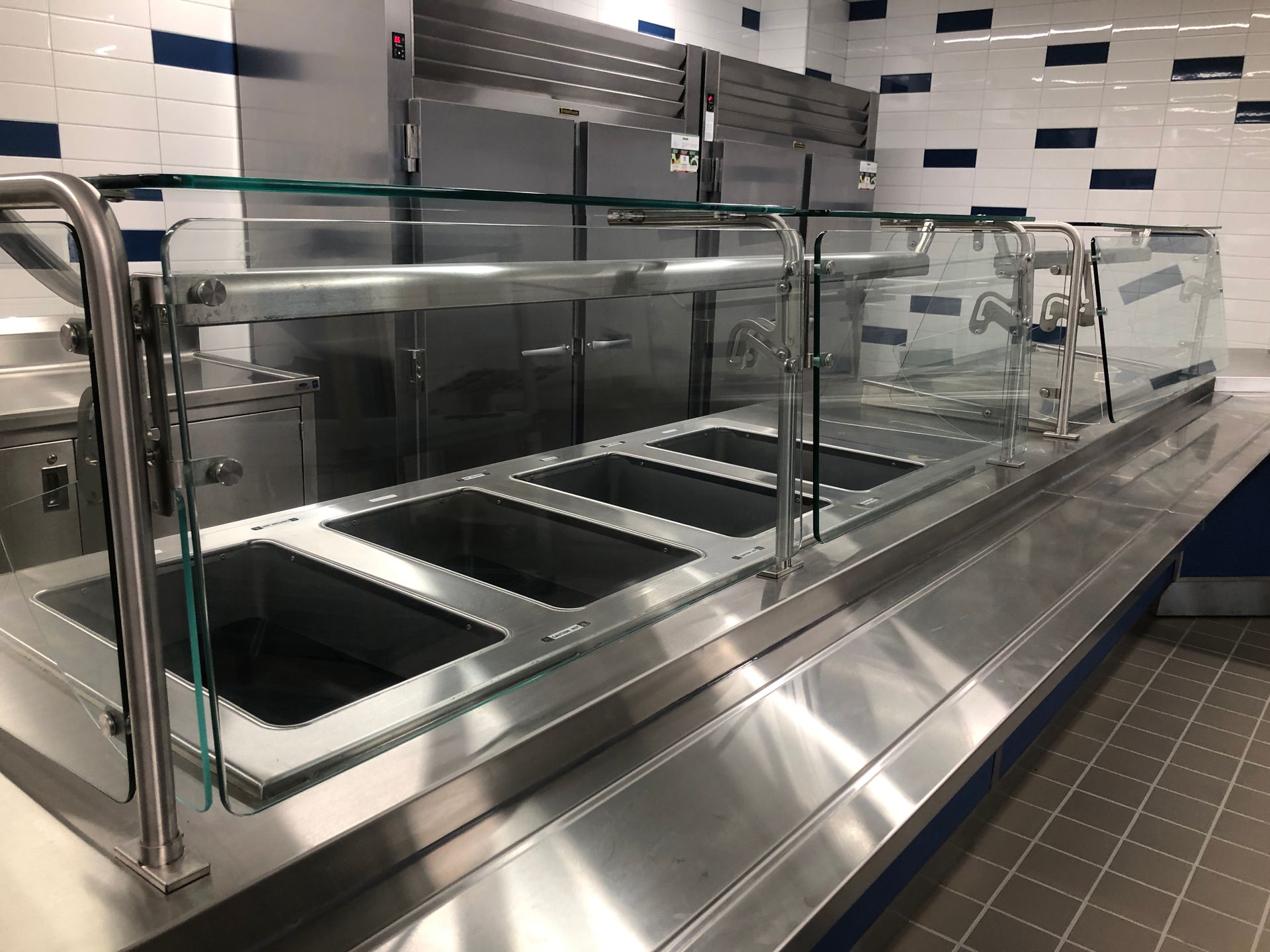 A stainless steel commercial food serving station with four recessed wells and a protective glass sneeze guard.