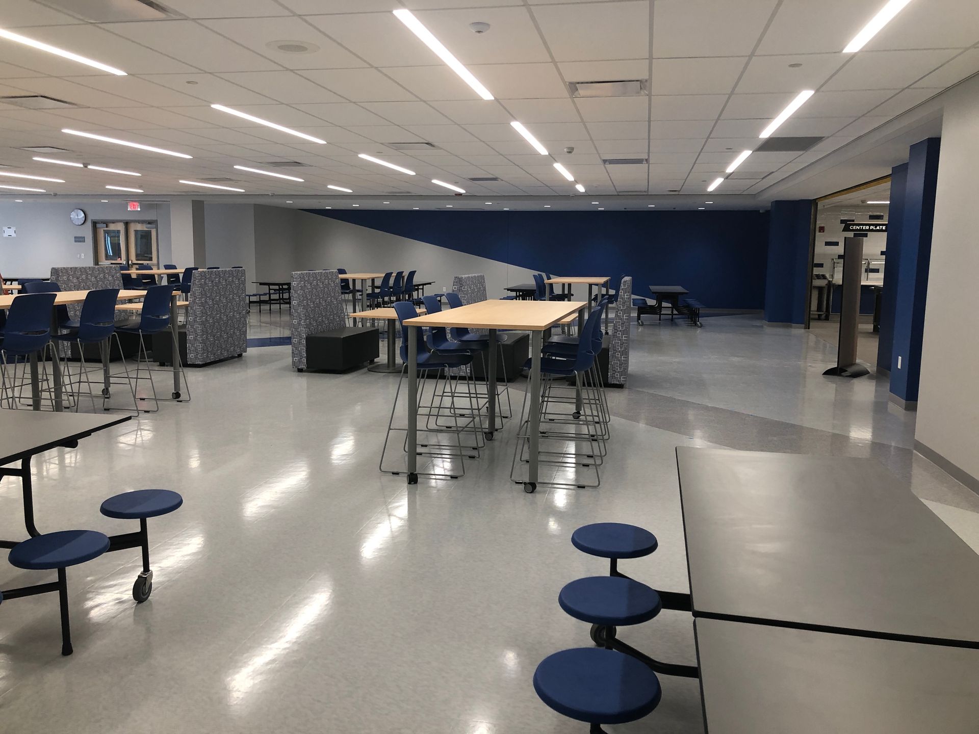 A brightly lit school cafeteria featuring rows of tables with attached blue stools and tall tables with chairs.