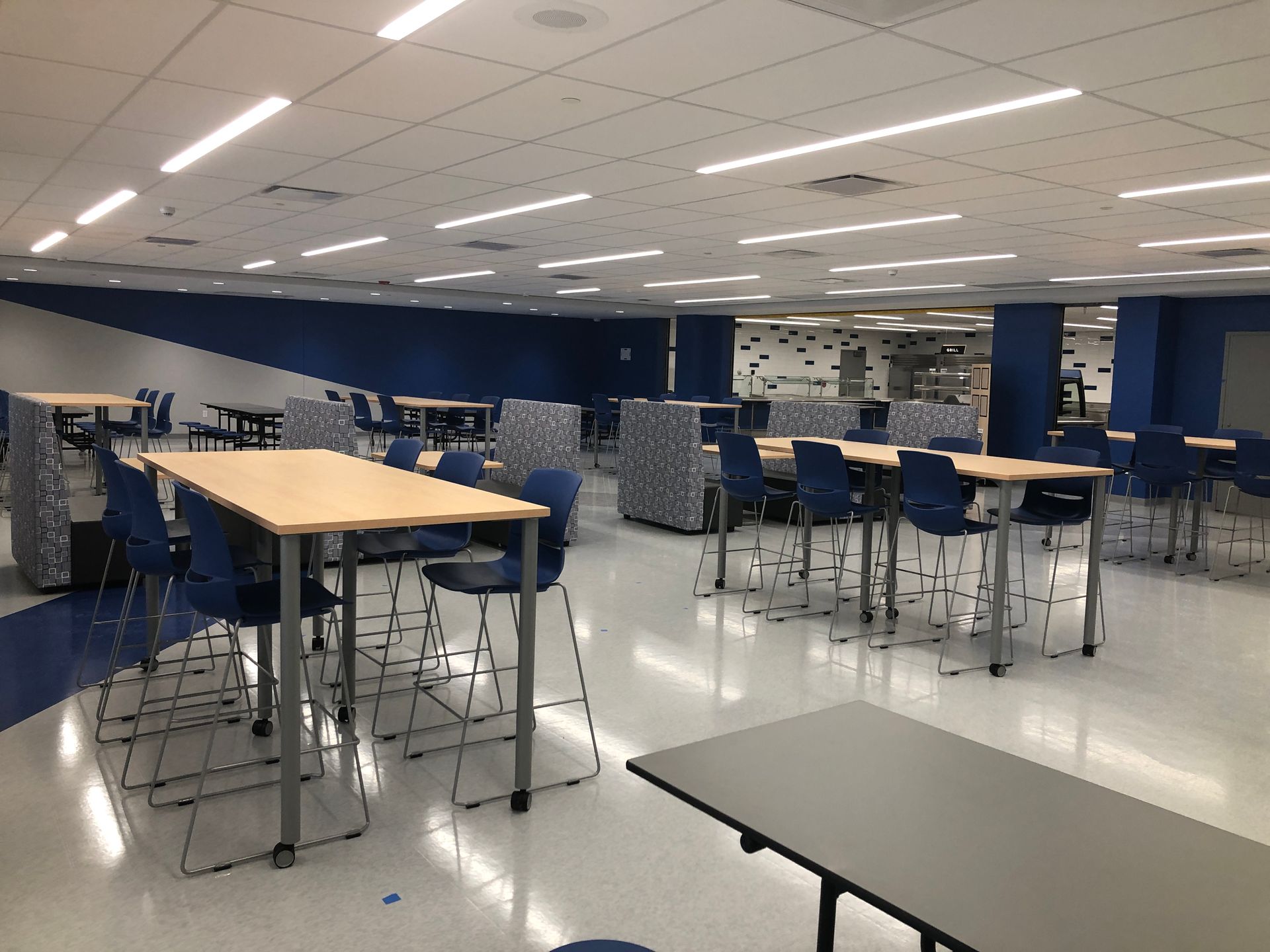 A brightly lit modern school cafeteria or break room with blue accents, tall tables, and chairs on a light-colored floor.