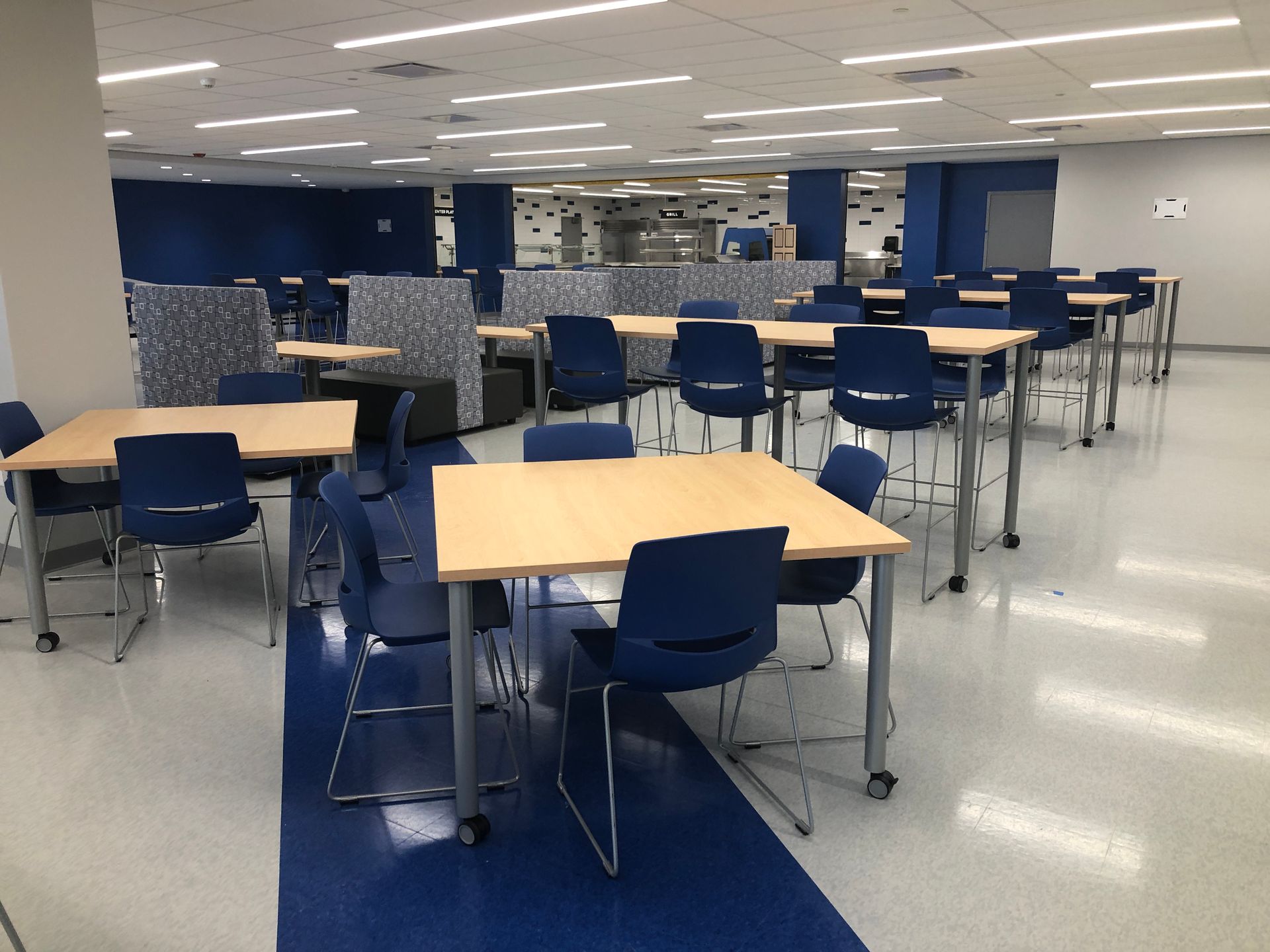 A brightly lit, modern office or study space with light wood tables, blue chairs, and blue accents on the floors and walls.