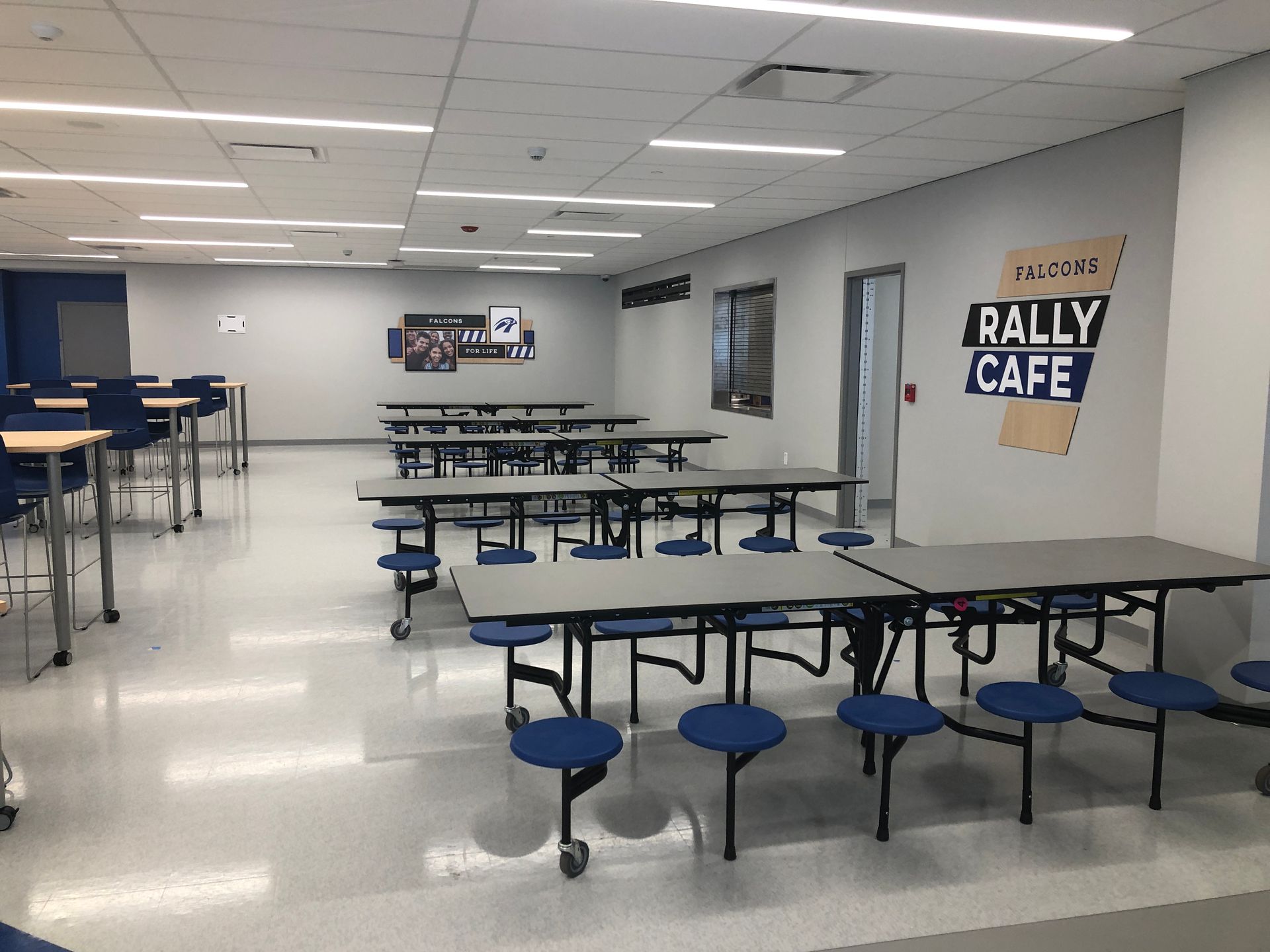 A brightly lit school cafeteria with rows of foldable tables and blue circular stools, featuring a 