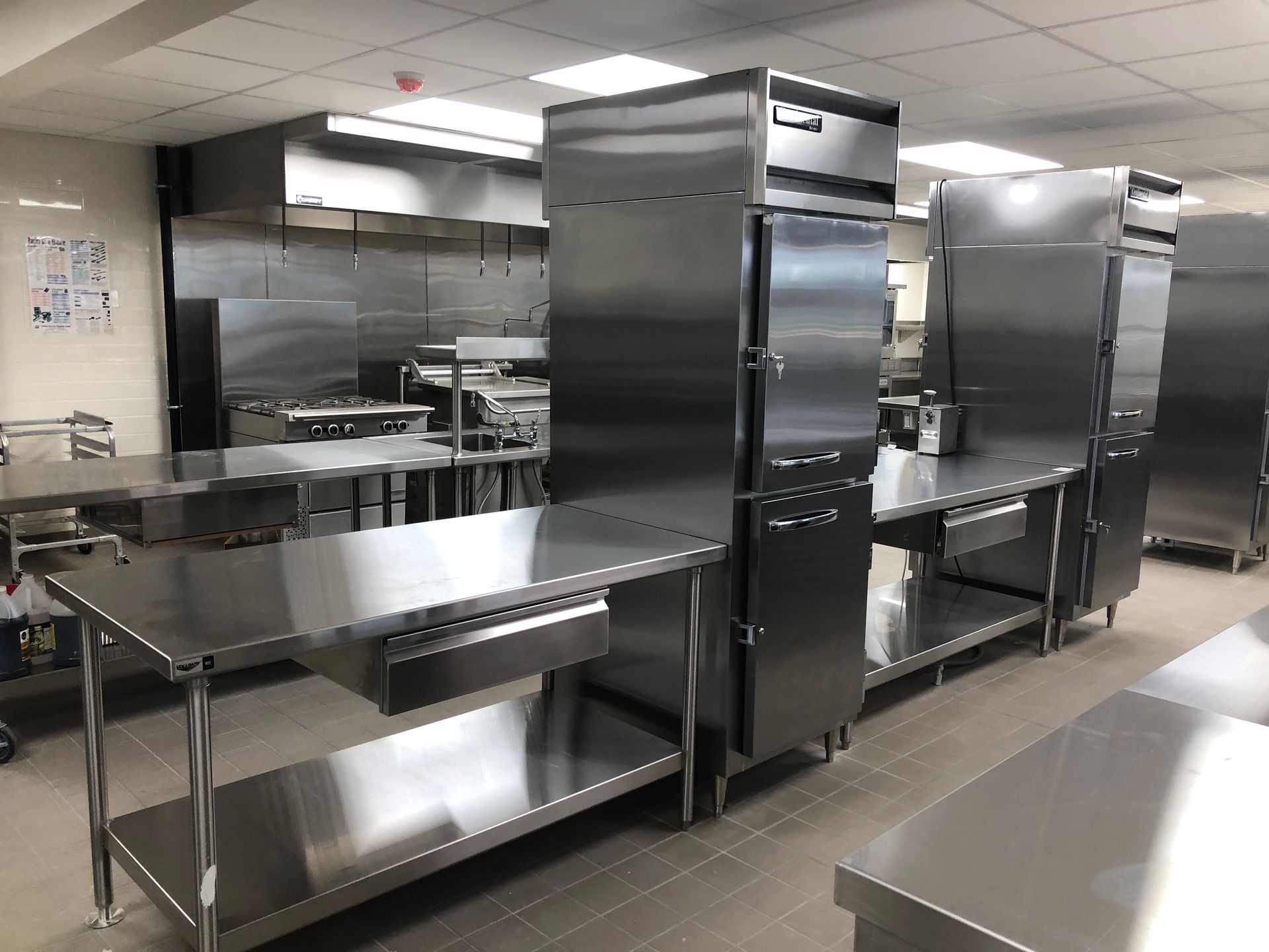 A clean, modern commercial kitchen with stainless steel prep tables, upright refrigerators, and a ventilation hood.