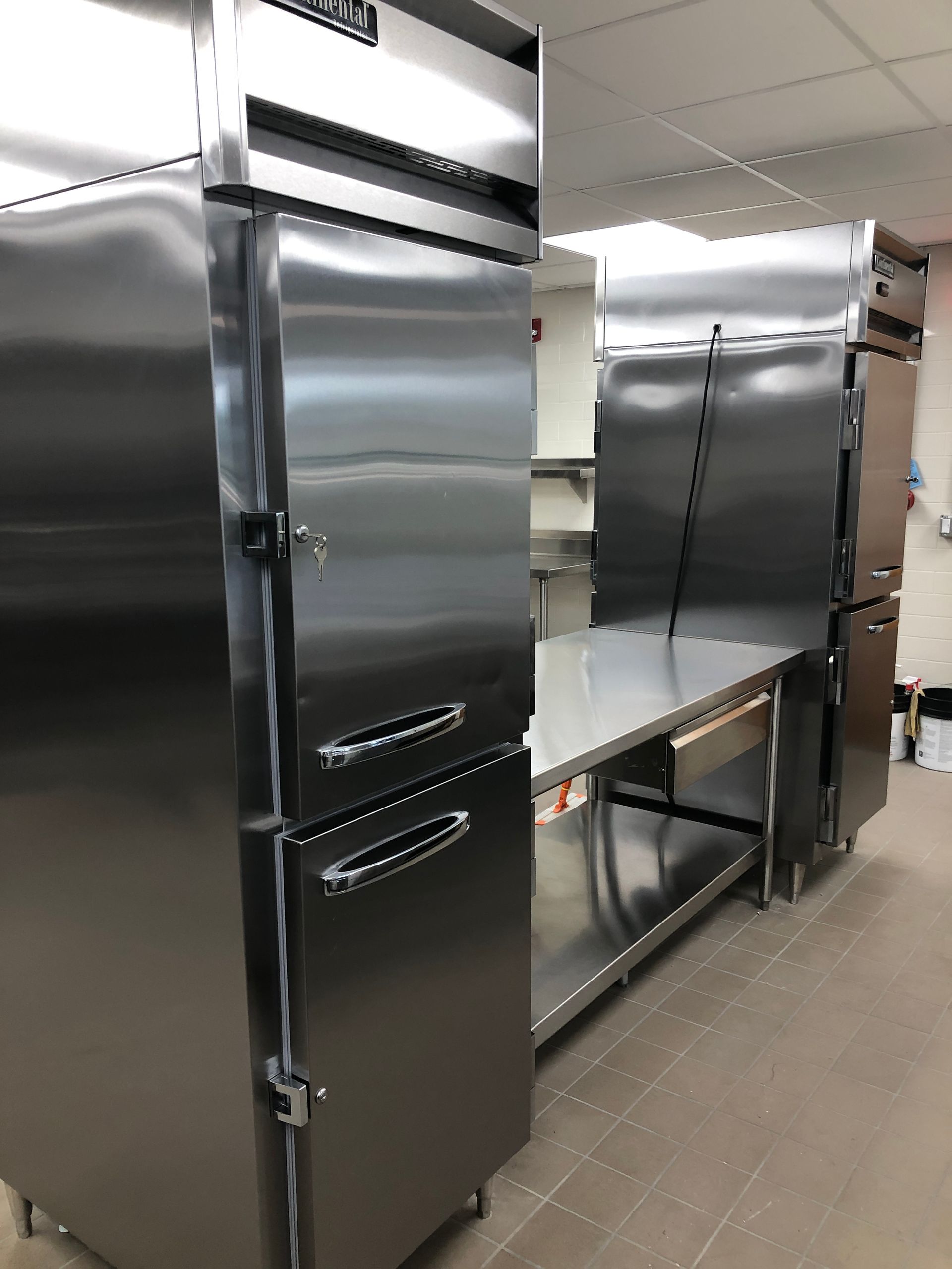 Two stainless steel industrial kitchen refrigerators flanking a stainless steel prep table in a commercial kitchen.