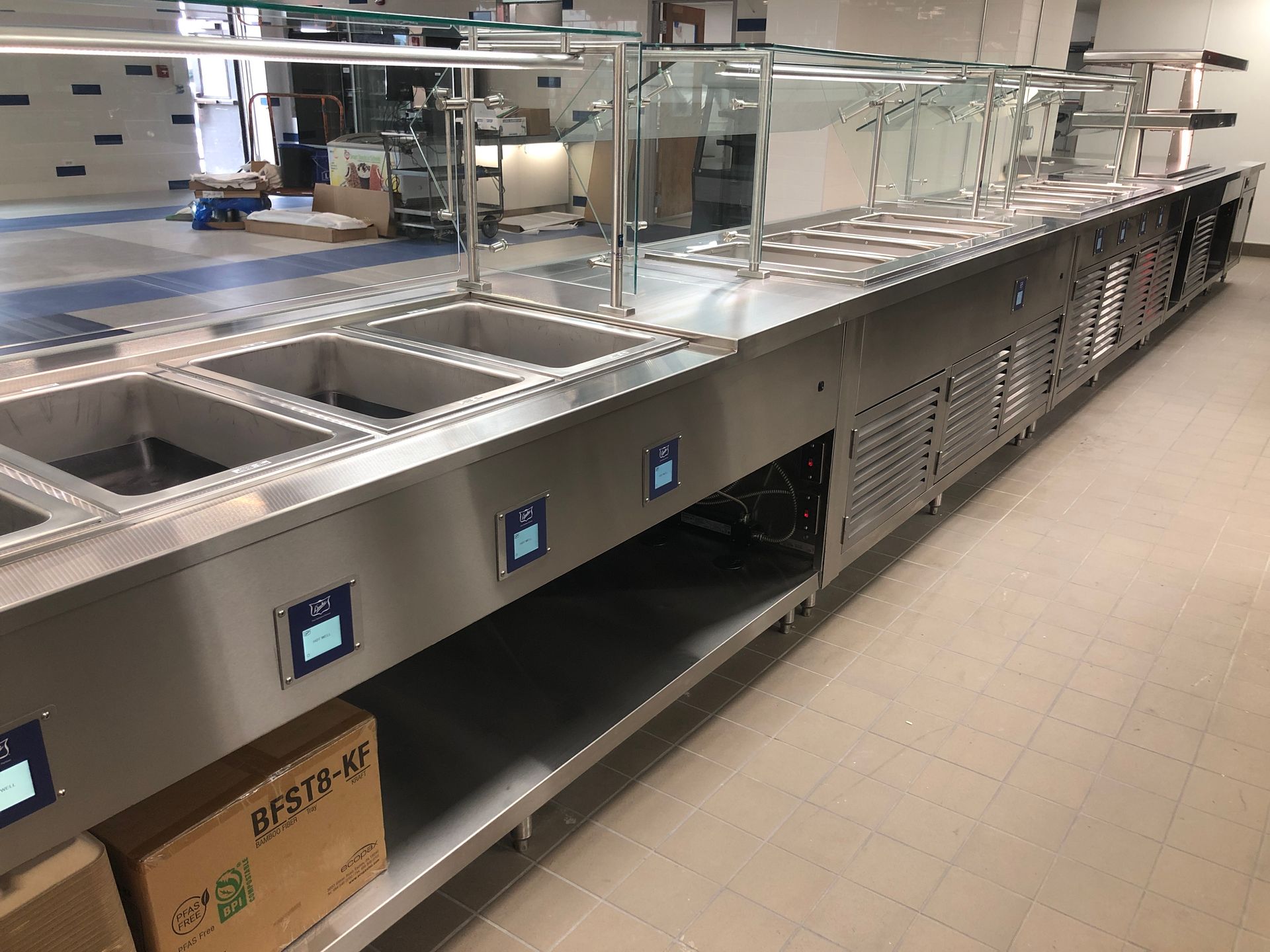 A long, stainless-steel food service buffet counter with multiple recessed basins, digital displays, and glass sneeze guards.