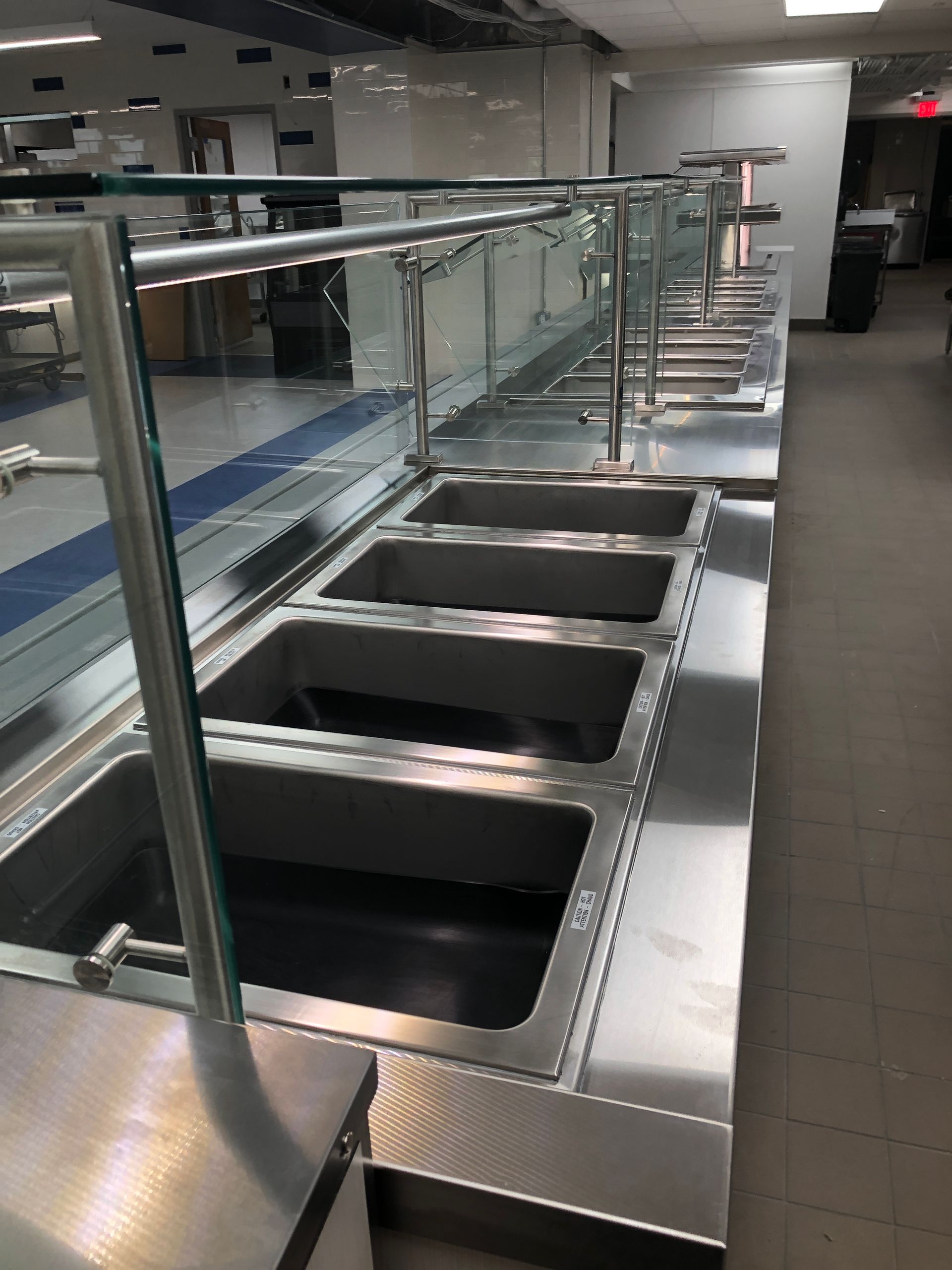 A stainless steel cafeteria serving counter with multiple empty food wells and a glass sneeze guard in an indoor space.