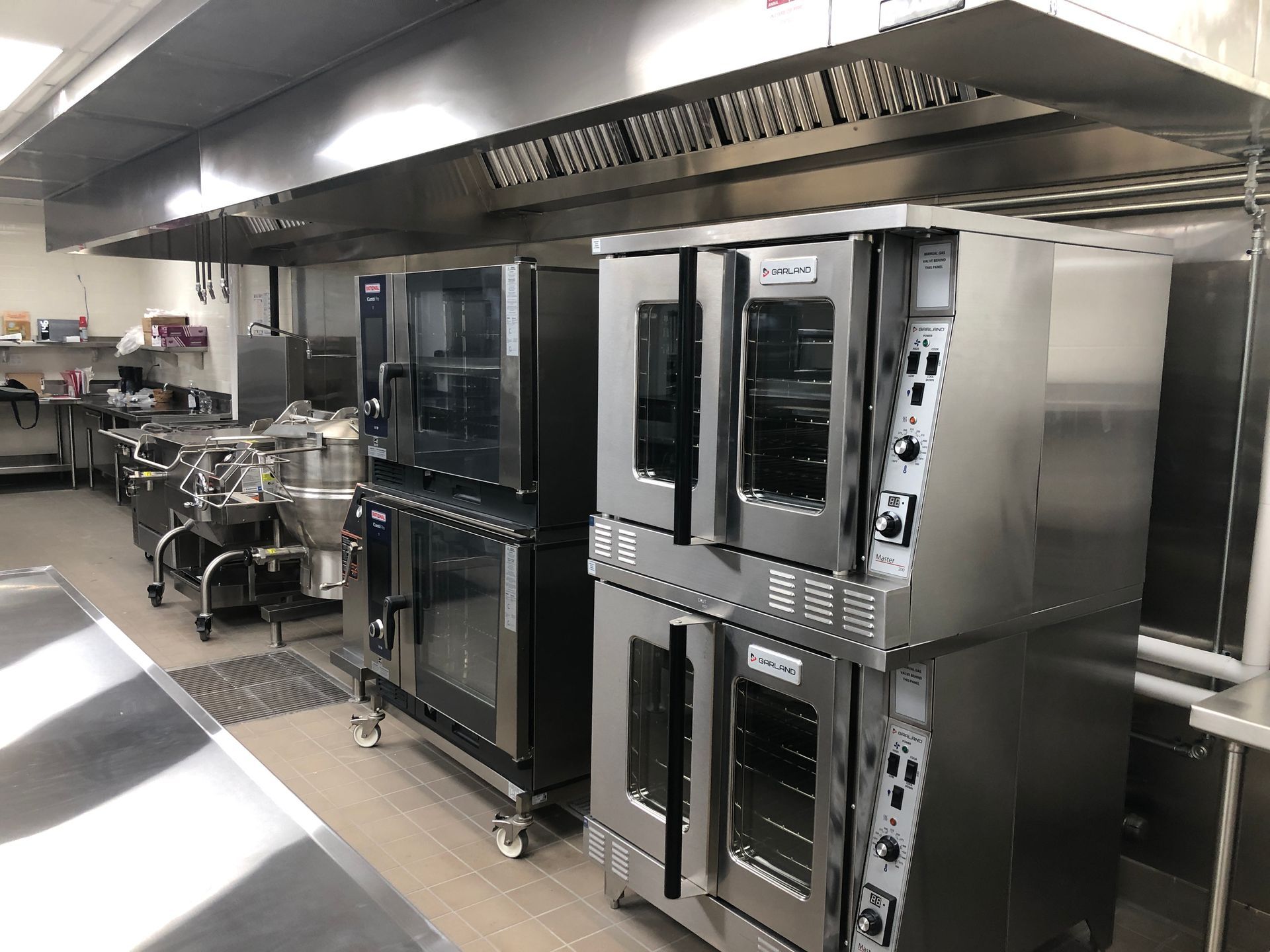 A row of stainless steel commercial ovens sits under a metallic ventilation hood in a professional kitchen.