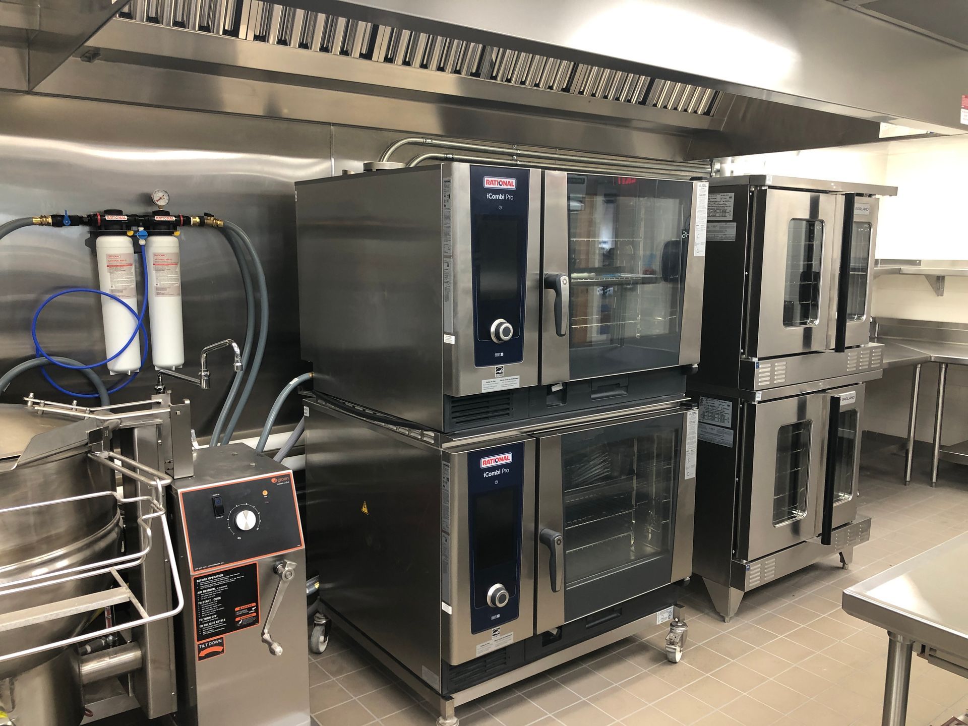 A commercial kitchen featuring stacked Rational combi ovens and a water filtration system under a stainless steel vent.
