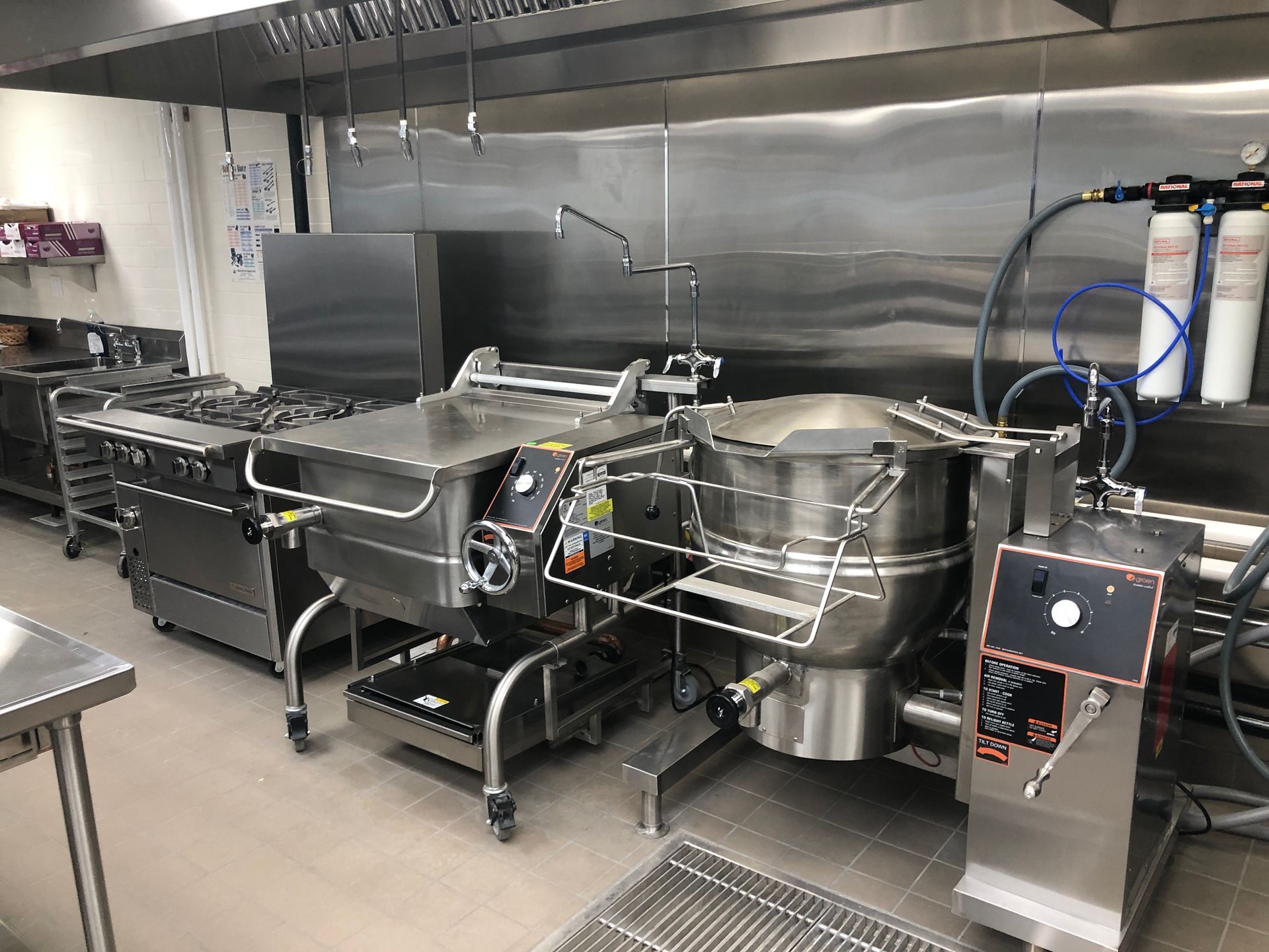 A stainless steel commercial kitchen featuring a tilting skillet, a large industrial steam kettle, and a gas stove.