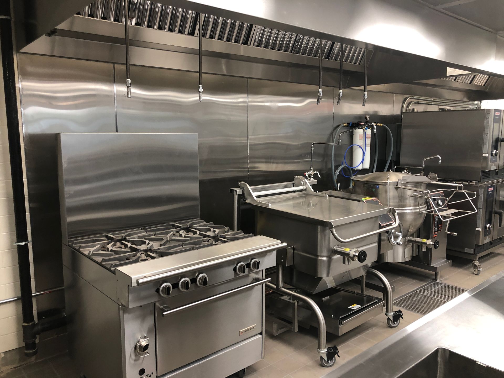 Professional stainless steel commercial kitchen equipment, including a range, tilting skillet, and kettle under a hood.
