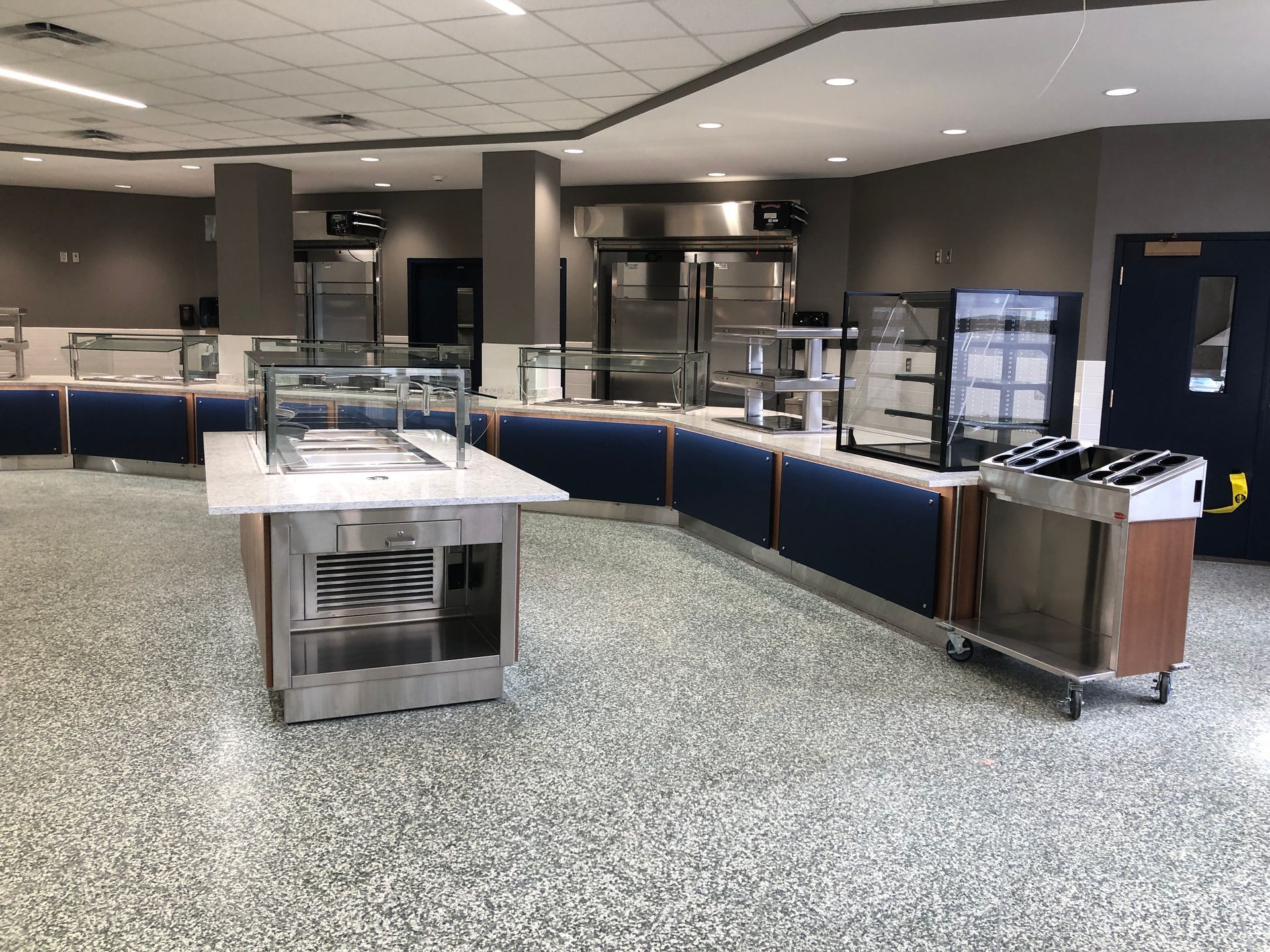 An empty school or cafeteria serving line with stainless steel stations, blue base panels, and a speckled floor.