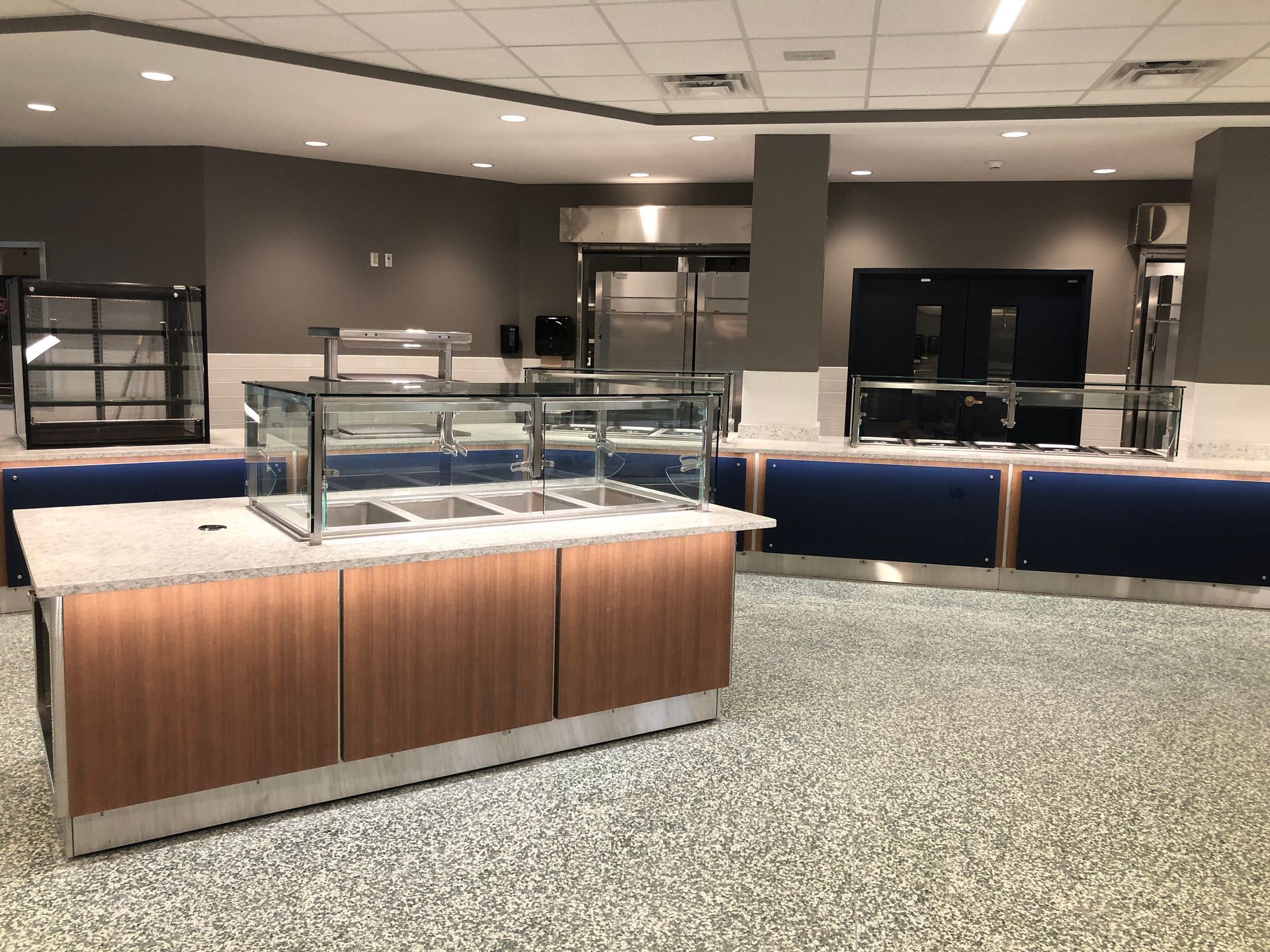 An empty cafeteria serving line with a wooden counter, a glass sneeze guard, and blue base panels on a speckled floor.