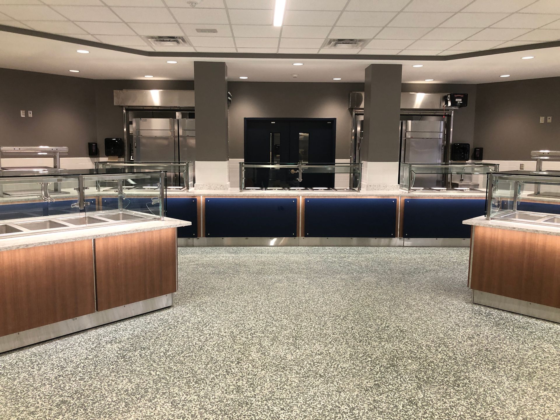 A buffet serving station with food warmers, wood-paneled counters, and blue accents in a modern, well-lit cafeteria.