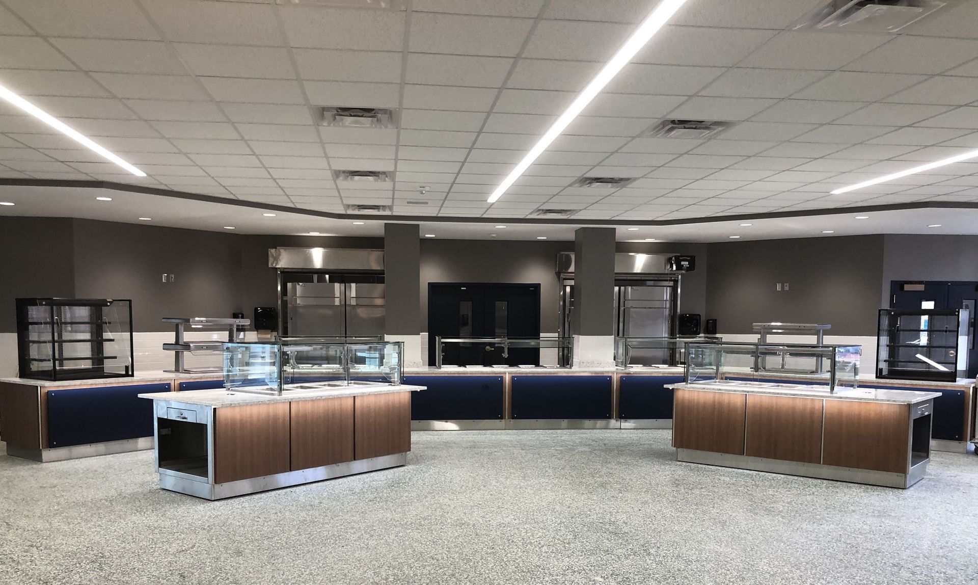 A modern cafeteria serving area with empty, glass-encased food stations featuring wood paneling, dark blue bases, and tiles.