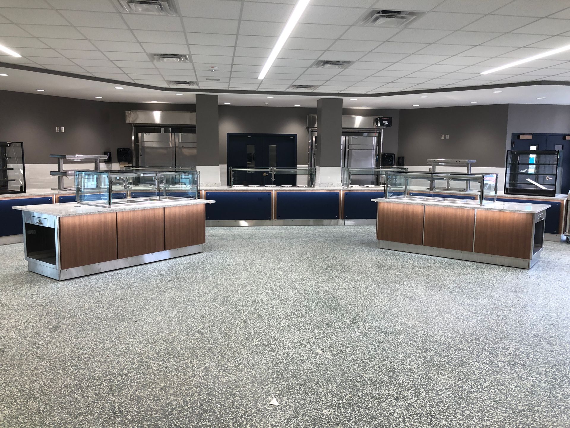 A modern, empty cafeteria with two wood-paneled serving stations, glass sneeze guards, and a gray speckled floor.