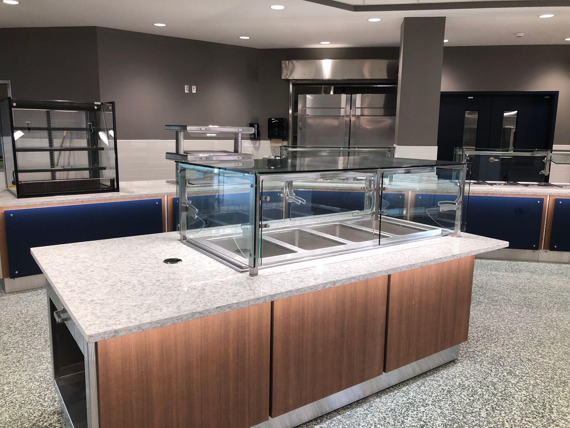 A modern cafeteria serving station with a granite countertop, glass sneeze guard, and wooden cabinetry in a large room.