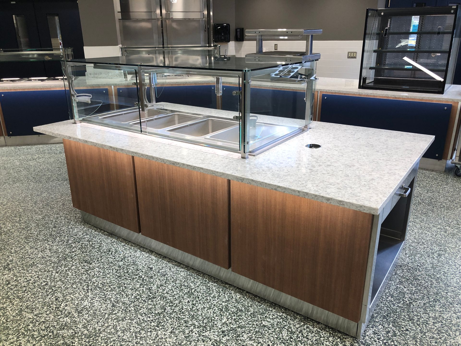 A commercial food service island with a quartz countertop, wood-paneled cabinets, and a metal sneeze guard in a cafeteria.