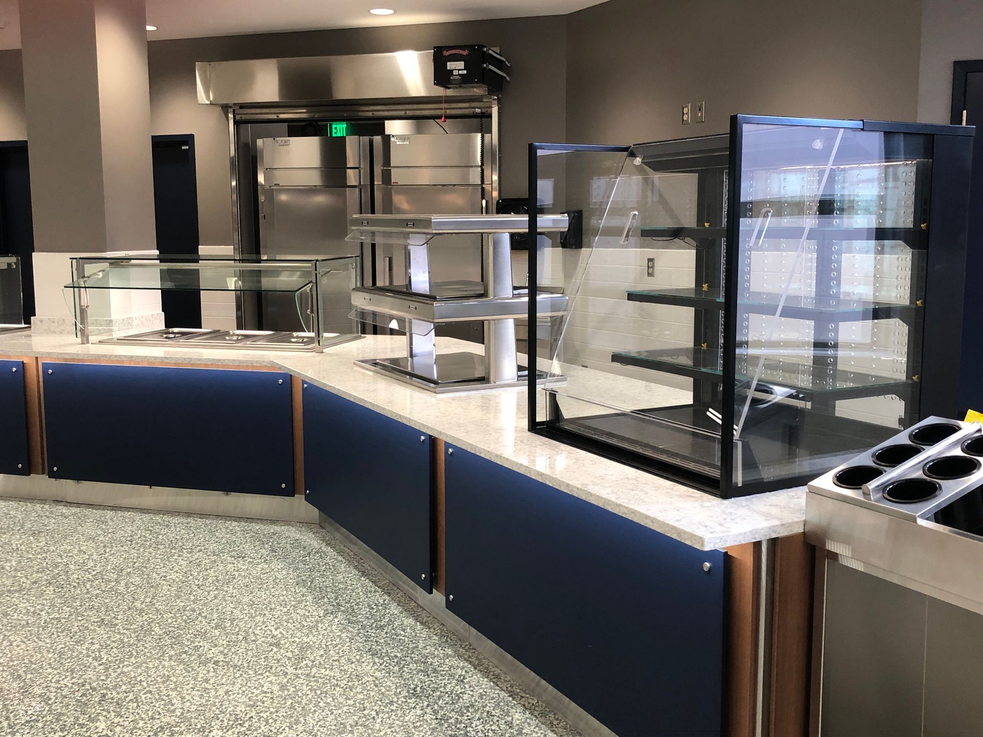 Empty cafeteria serving line with a glass food display case, stainless steel counters, and blue panels on a speckled floor.