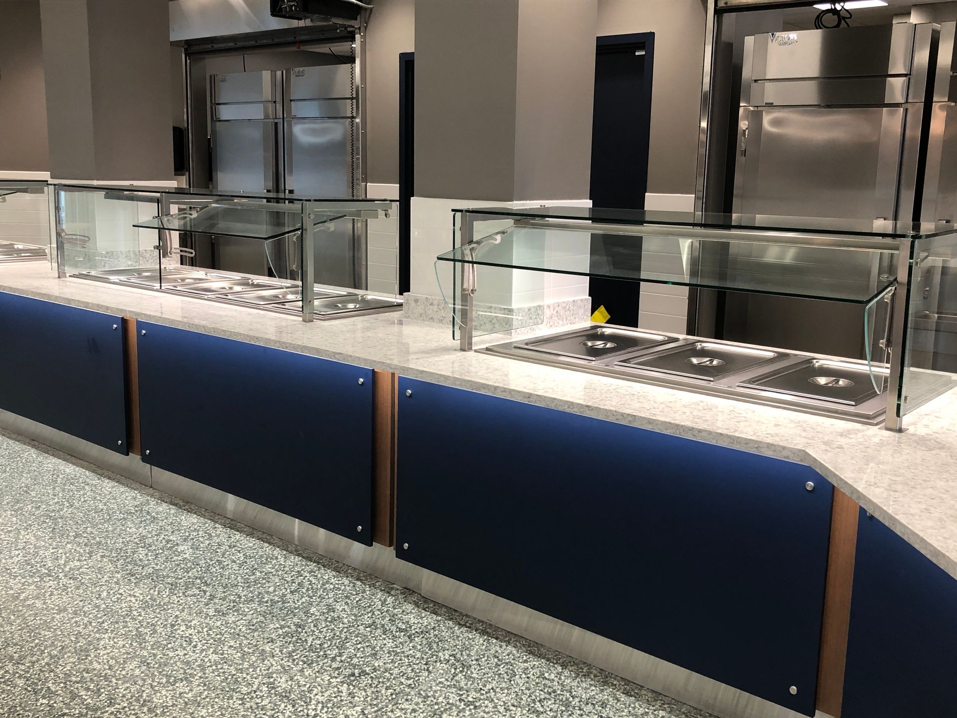 A buffet-style serving counter with blue panels, glass sneeze guards, and granite countertops in a modern dining hall.