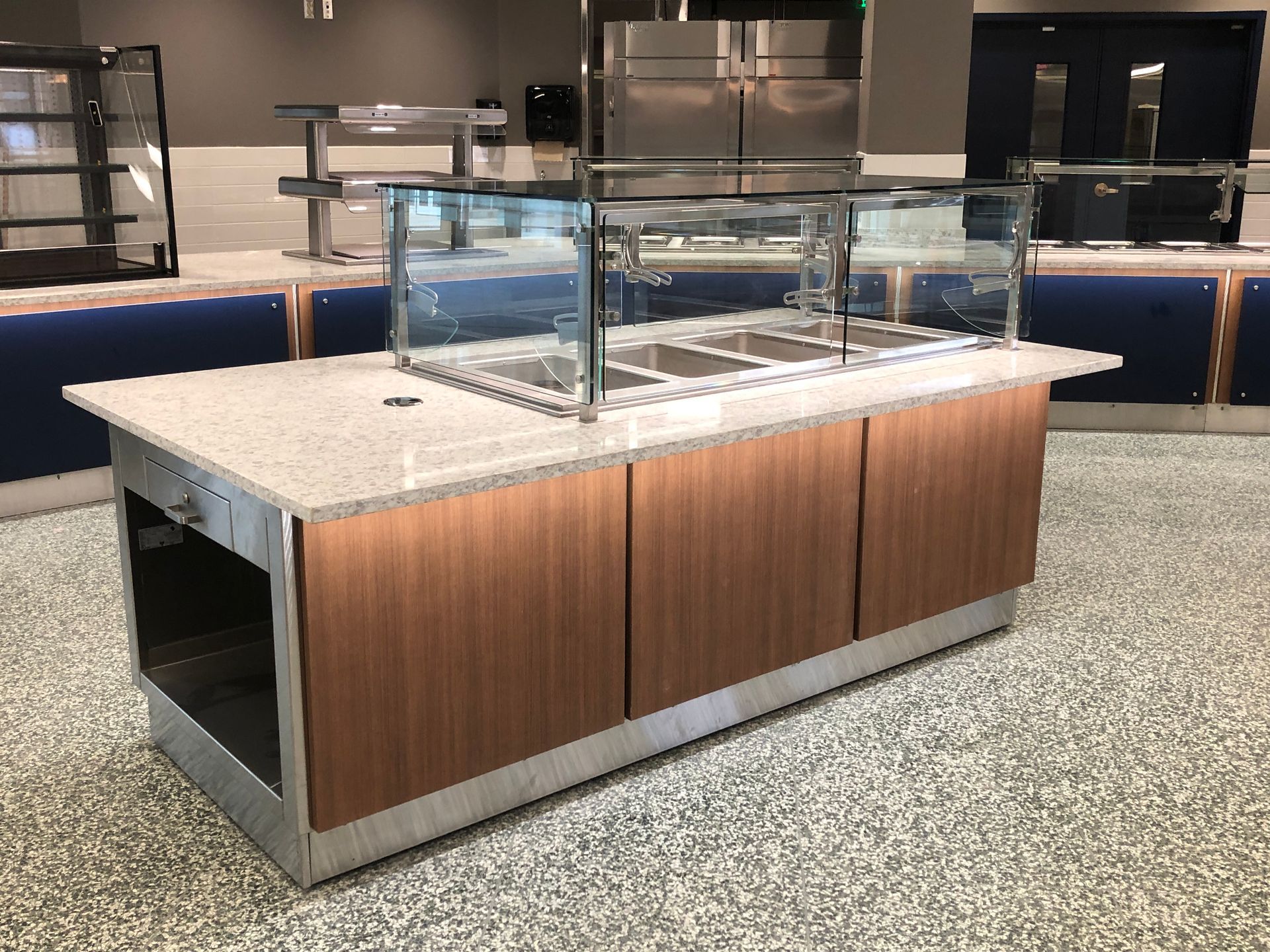 A modern salad bar station with a granite countertop, wood-paneled base, and protective glass sneeze guard in a kitchen.