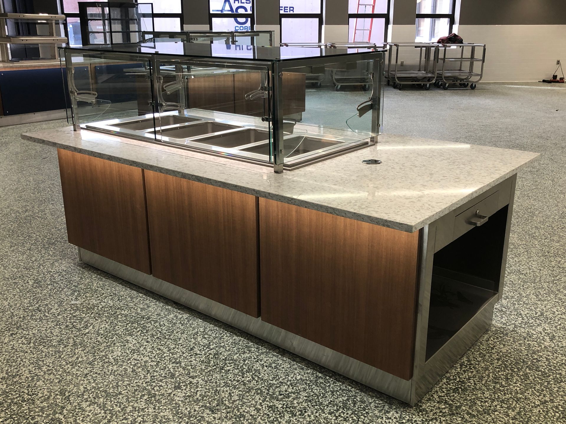 A commercial food service island with a quartz countertop, glass sneeze guard, and lit wood-paneled base on a stone floor.