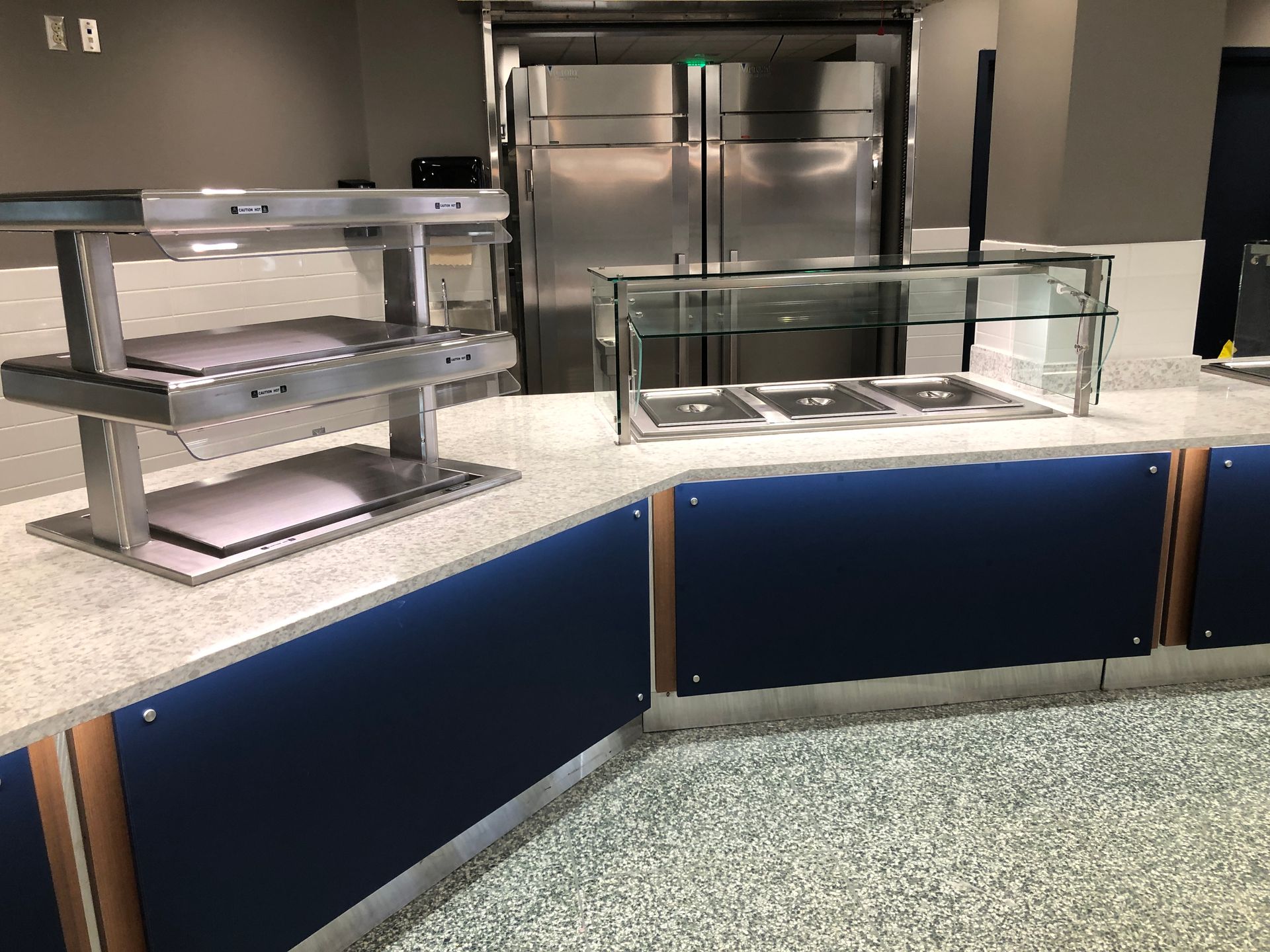 A modern food service station with a stainless steel sneeze guard, countertop, and blue panels in a dining hall setting.