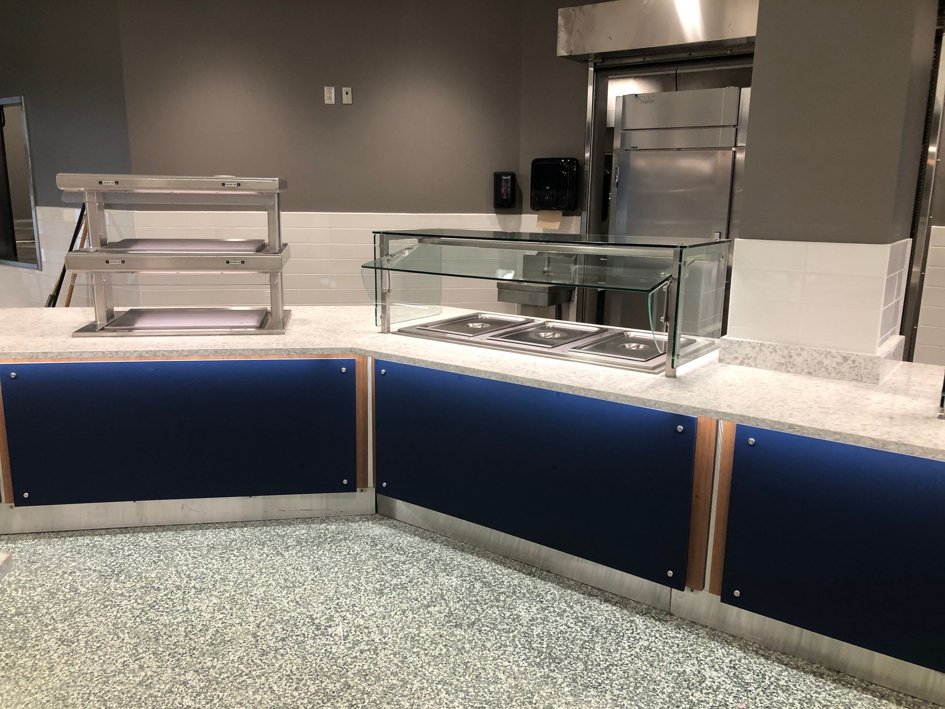 A service counter with a stainless steel food warmer and glass sneeze guard on a speckled countertop.