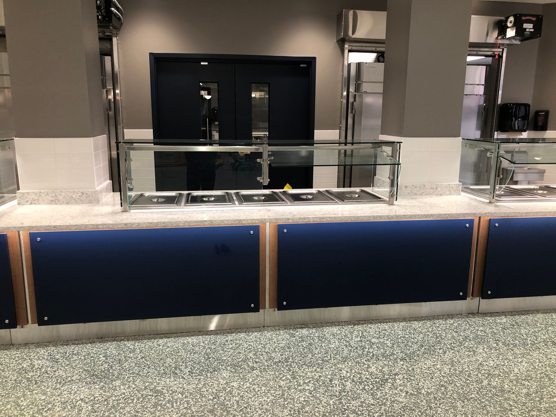 A buffet service counter with empty metal food wells, glass sneeze guards, and blue panels in a neutral-toned room.