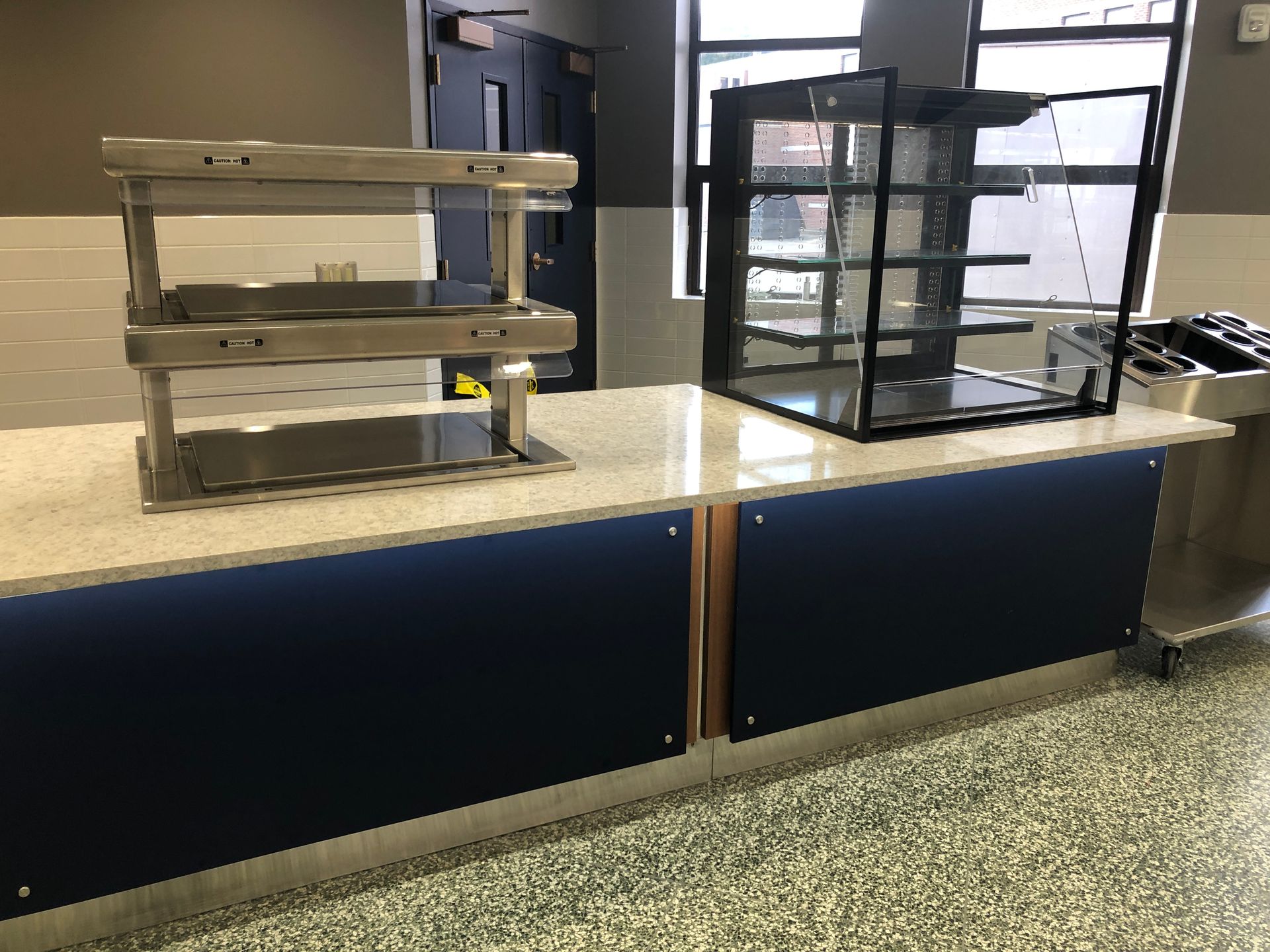 A stainless steel countertop with a food warming rack and a glass display case inside a brightly lit cafeteria.